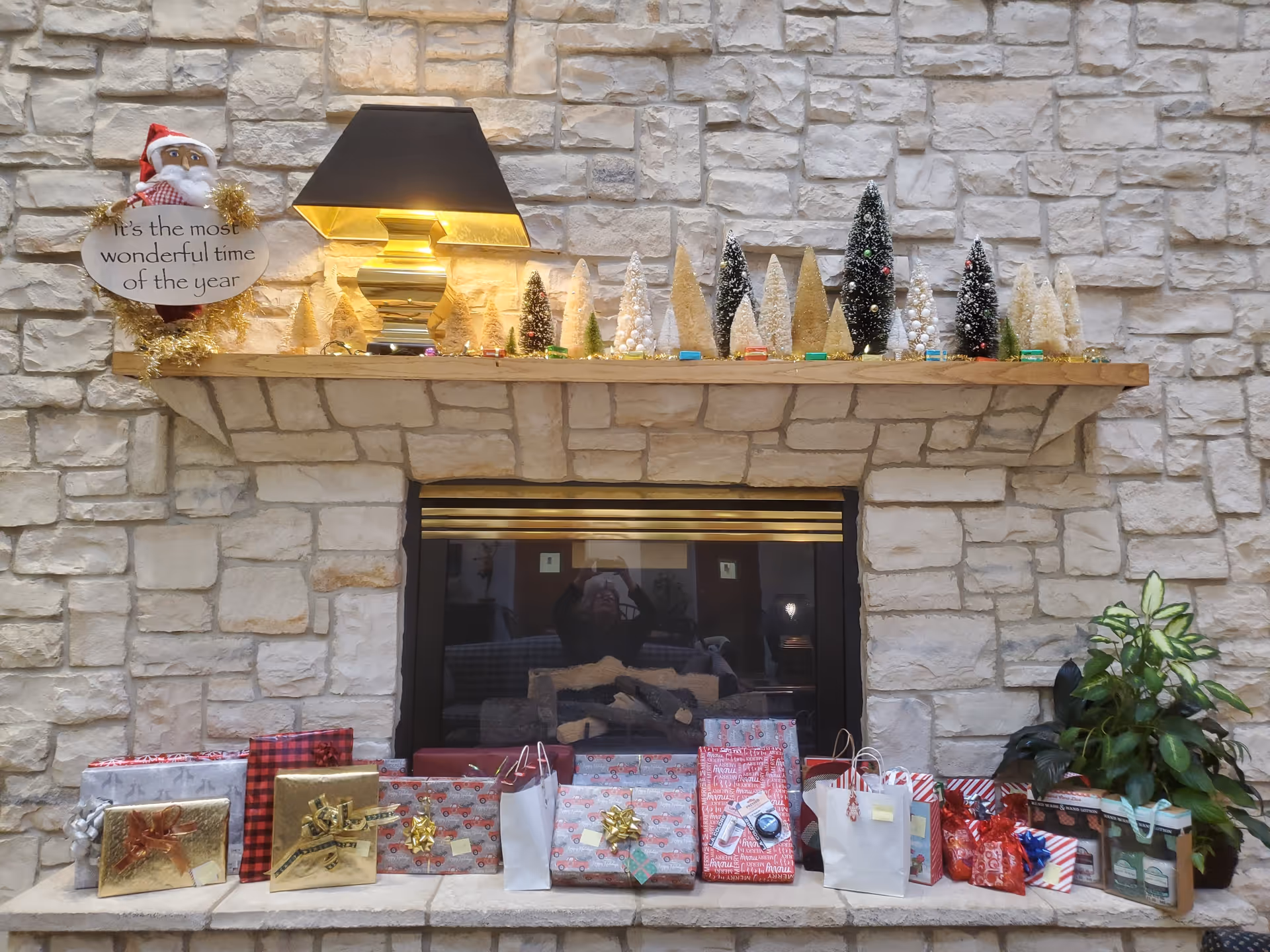 Stone fireplace mantel decorated with small Christmas trees and a lamp, with wrapped presents on the hearth and a potted plant to the right.