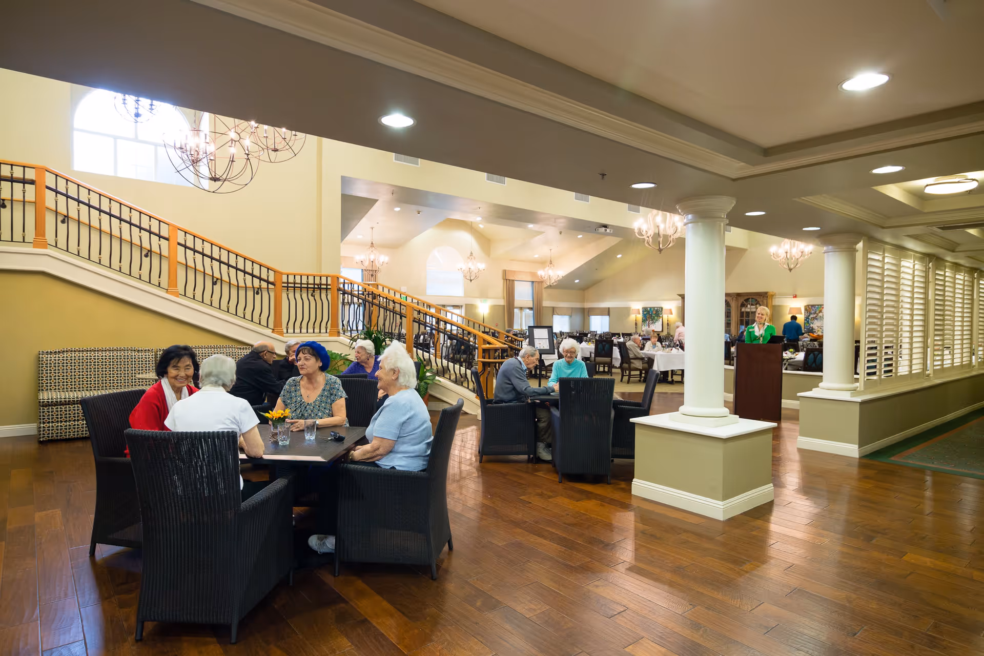 A group of elderly people sitting and chatting around tables in a spacious, well-lit common area with wooden floors, chandeliers, and a staircase with a wooden handrail in the background. The room has large windows and white columns, creating a warm and inviting atmosphere.