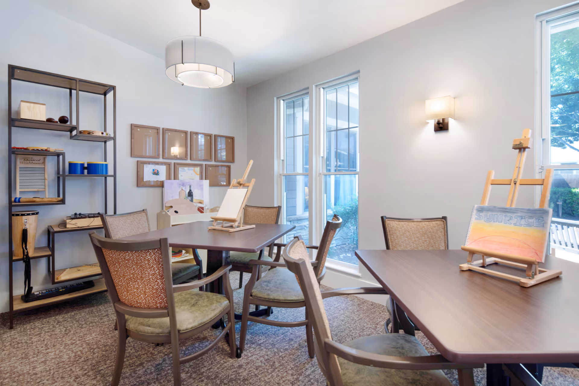 A bright activity room with two wooden tables and several cushioned chairs arranged around them. On one table, there is a small easel holding a drawing with the word 'SUNRISE' and a sunset illustration. The other table has a blank easel. The room has large windows letting in natural light, a modern ceiling light fixture, a wall sconce, and a metal shelving unit with various musical instruments and art supplies. Several framed artworks are displayed on the wall.