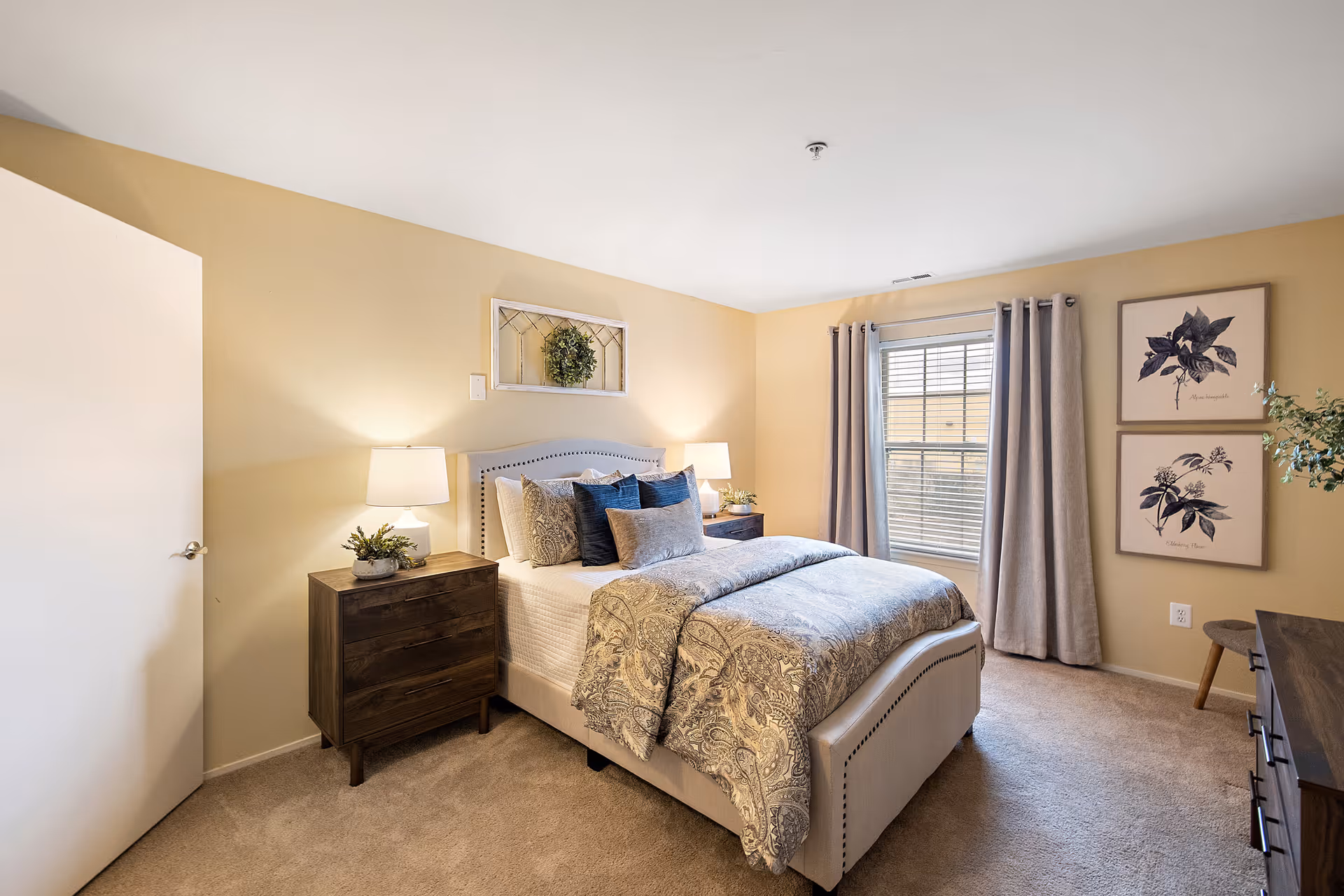 A cozy bedroom with a neatly made bed featuring patterned bedding and multiple pillows. There are two wooden nightstands on either side of the bed, each with a lamp and small plant. A window with gray curtains lets in natural light. The walls are painted a soft beige, decorated with two framed botanical prints and a decorative mirror with a wreath above the bed. The room has beige carpeting and a closed white door on the left side.