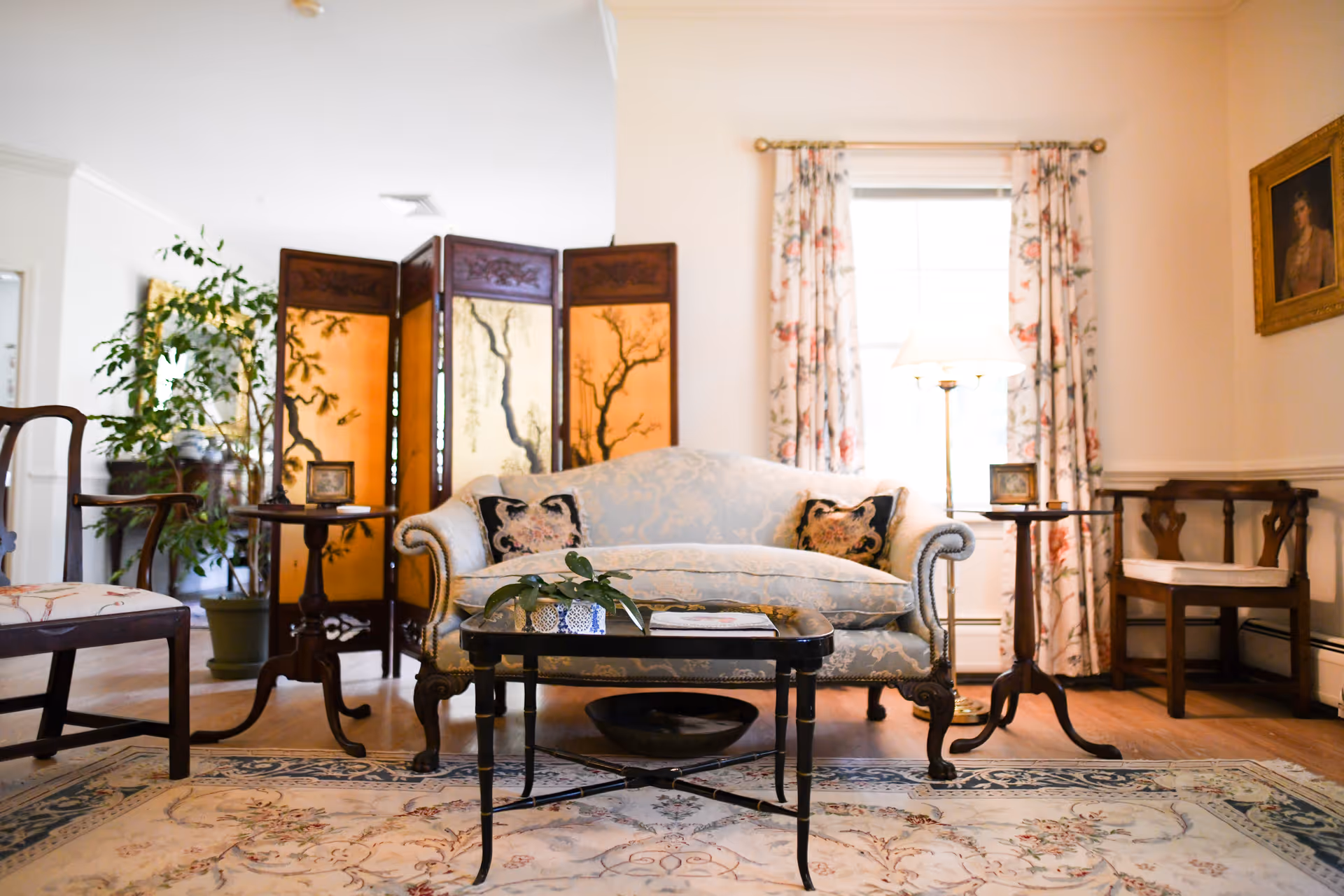 A cozy living room with a light blue upholstered sofa adorned with two decorative pillows, a black coffee table with a small plant on it, floral curtains framing a window, a wooden folding screen with artistic tree designs, and several wooden chairs. The room has a warm and inviting atmosphere with a patterned rug on the floor and a framed portrait on the wall.