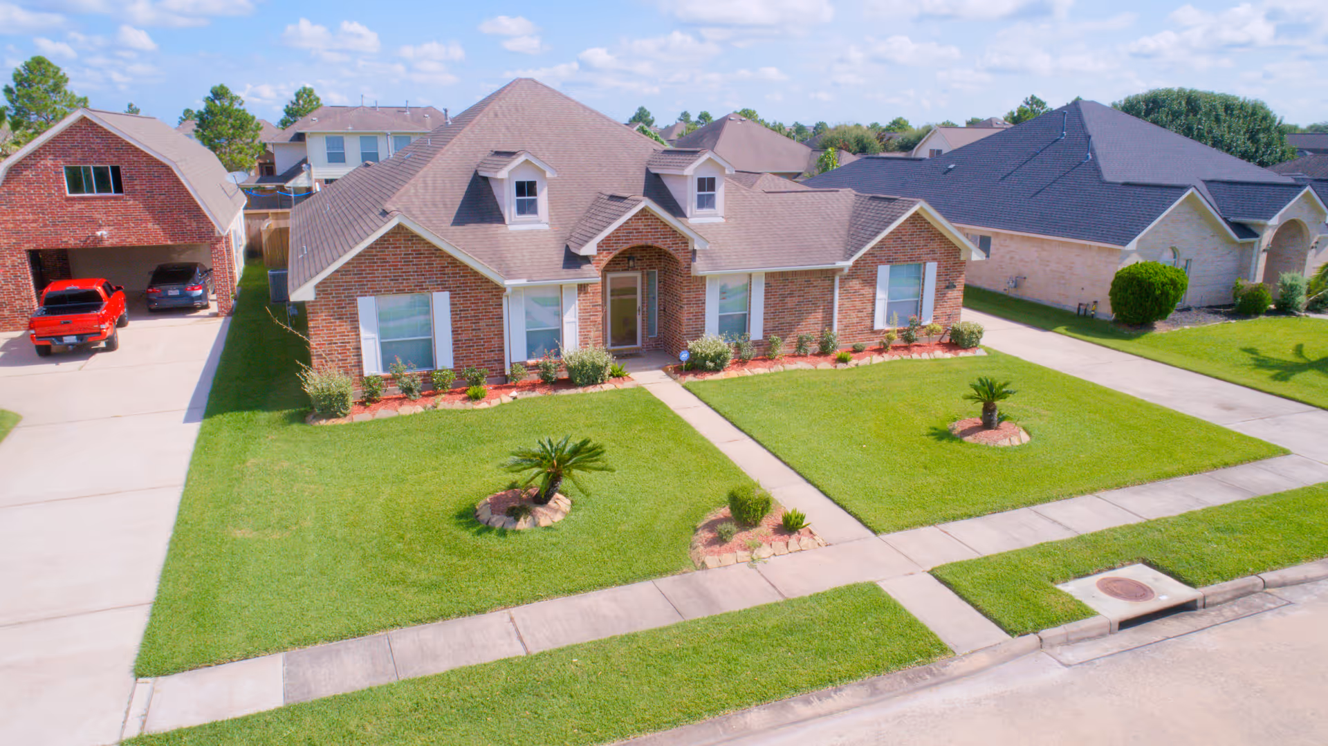 A single-story brick house with a well-maintained front lawn featuring small landscaped garden beds and a concrete walkway leading to the front door. The house has multiple windows with white shutters and a gabled roof. There is a driveway on the left side with two parked vehicles and neighboring houses visible on either side under a partly cloudy sky.