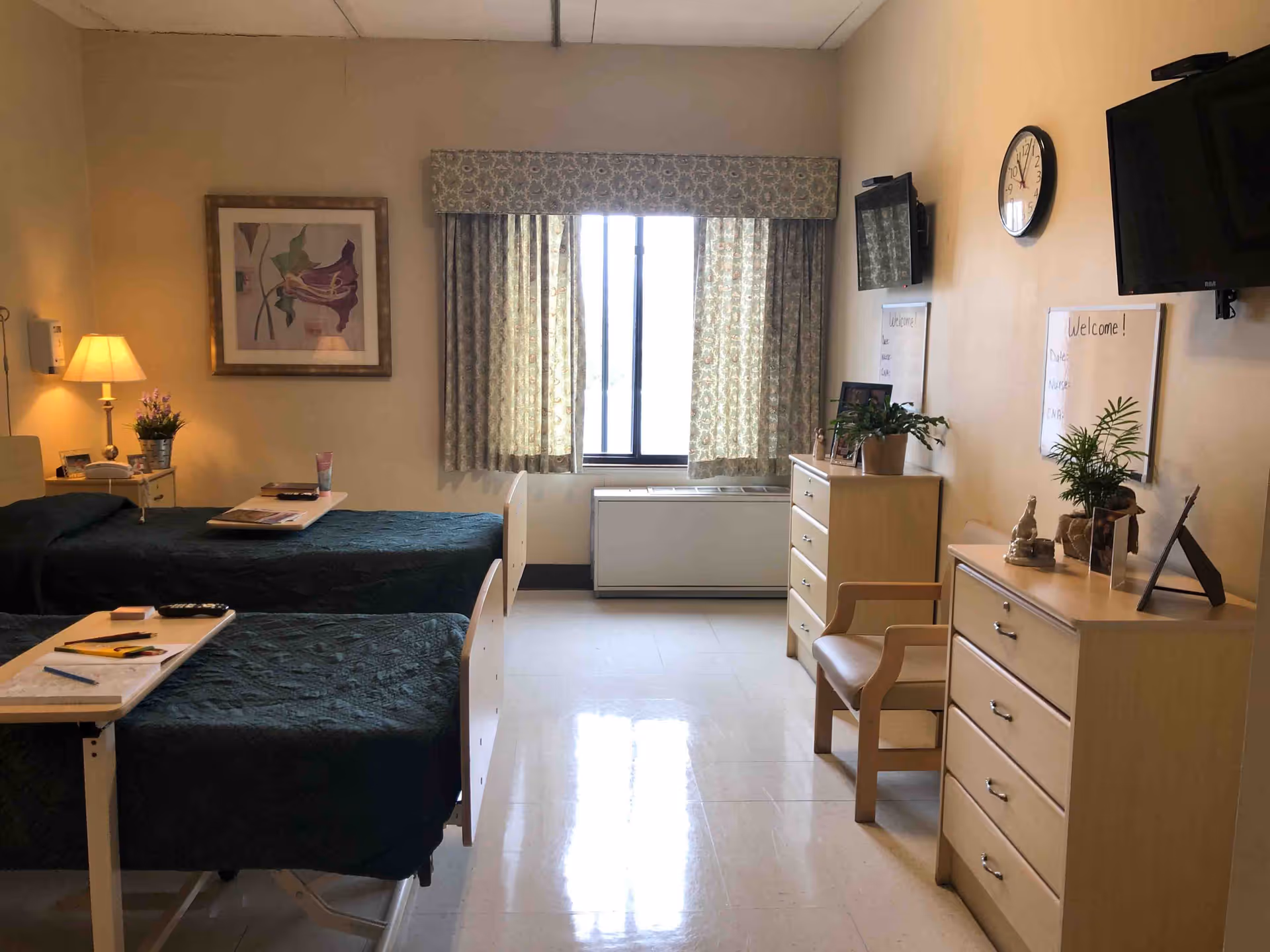 Shared senior facility bedroom with two single beds, dressers, a window with curtains, wall-mounted TV and a clock.