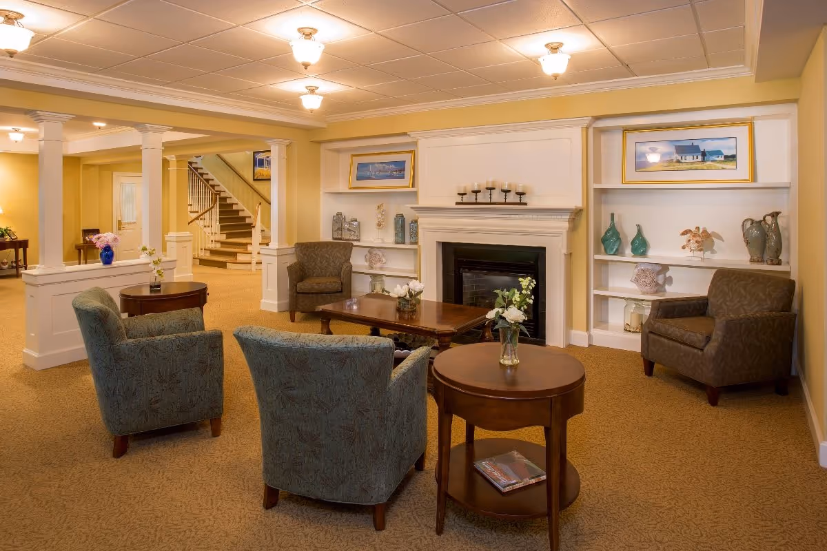 A cozy living room area in a senior living facility with four upholstered armchairs arranged around two wooden coffee tables. The room features a white fireplace with candles on the mantel, built-in shelves with decorative items and framed pictures, beige carpet, and soft yellow walls. In the background, there is a staircase and additional seating areas.
