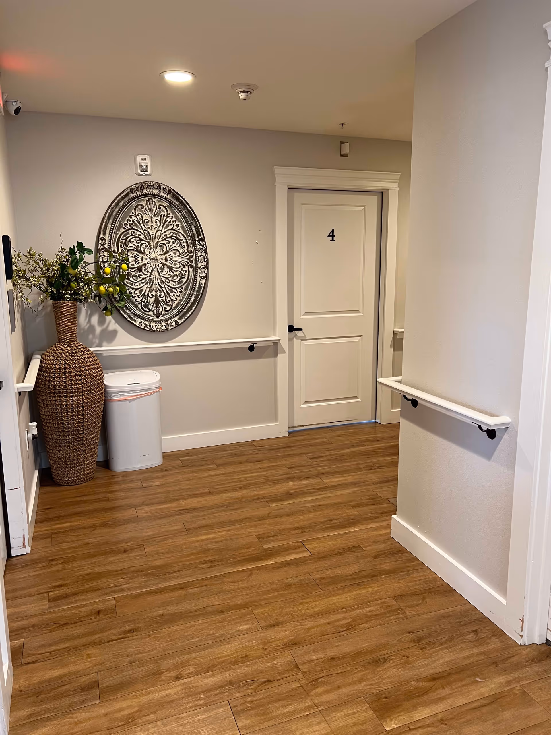 Interior hallway with wooden flooring, beige walls, and white trim. A white door labeled with the number 4 is visible at the end of the hallway. On the left side, there is a large woven vase with green and yellow artificial plants, a white trash can, and a decorative round wall hanging with an intricate pattern. Handrails are mounted on both sides of the hallway walls.