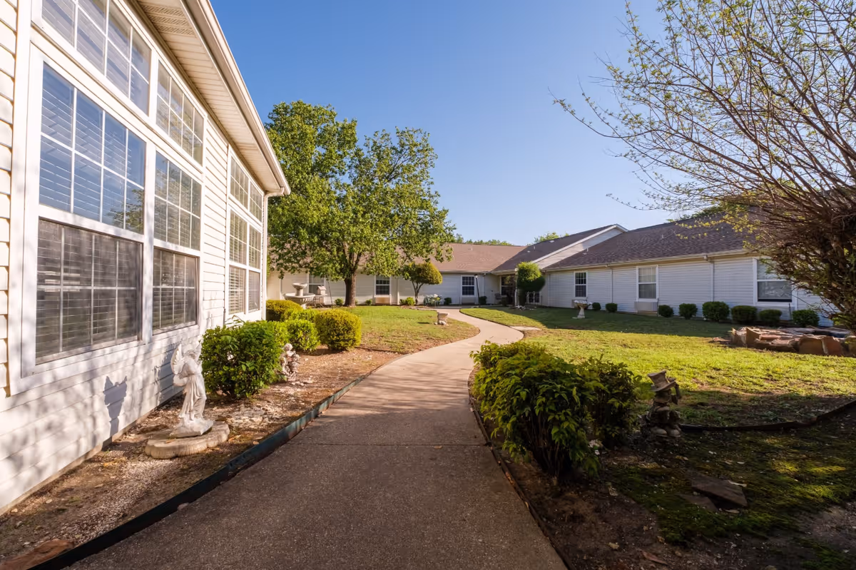 Sunlit courtyard walkway between single-story senior living buildings with shrubs, trees, and small garden statues.