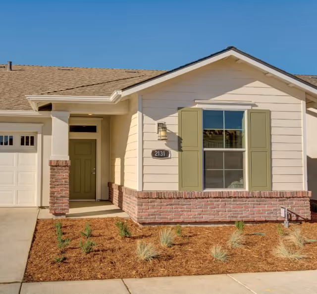 Front exterior view of a single-story house with beige siding, green shutters, a green front door, a brick accent wall, and a small landscaped area with young plants and mulch under a clear blue sky.