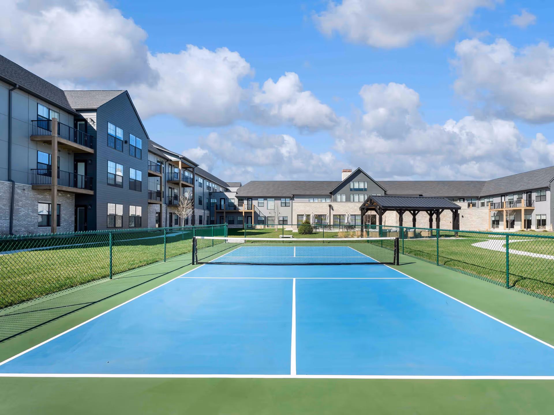 Outdoor tennis court with a blue playing surface and green borders, surrounded by a green chain-link fence. In the background, there is a large senior living facility building with multiple windows, balconies, and a covered pavilion. The sky is partly cloudy with patches of blue.