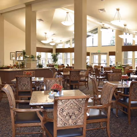 A spacious dining room with multiple tables and chairs arranged neatly. The room features large windows allowing natural light to fill the space, decorative light fixtures hanging from the ceiling, and floral centerpieces on the tables. The overall decor is warm and inviting with patterned upholstery on the chairs and a carpeted floor.