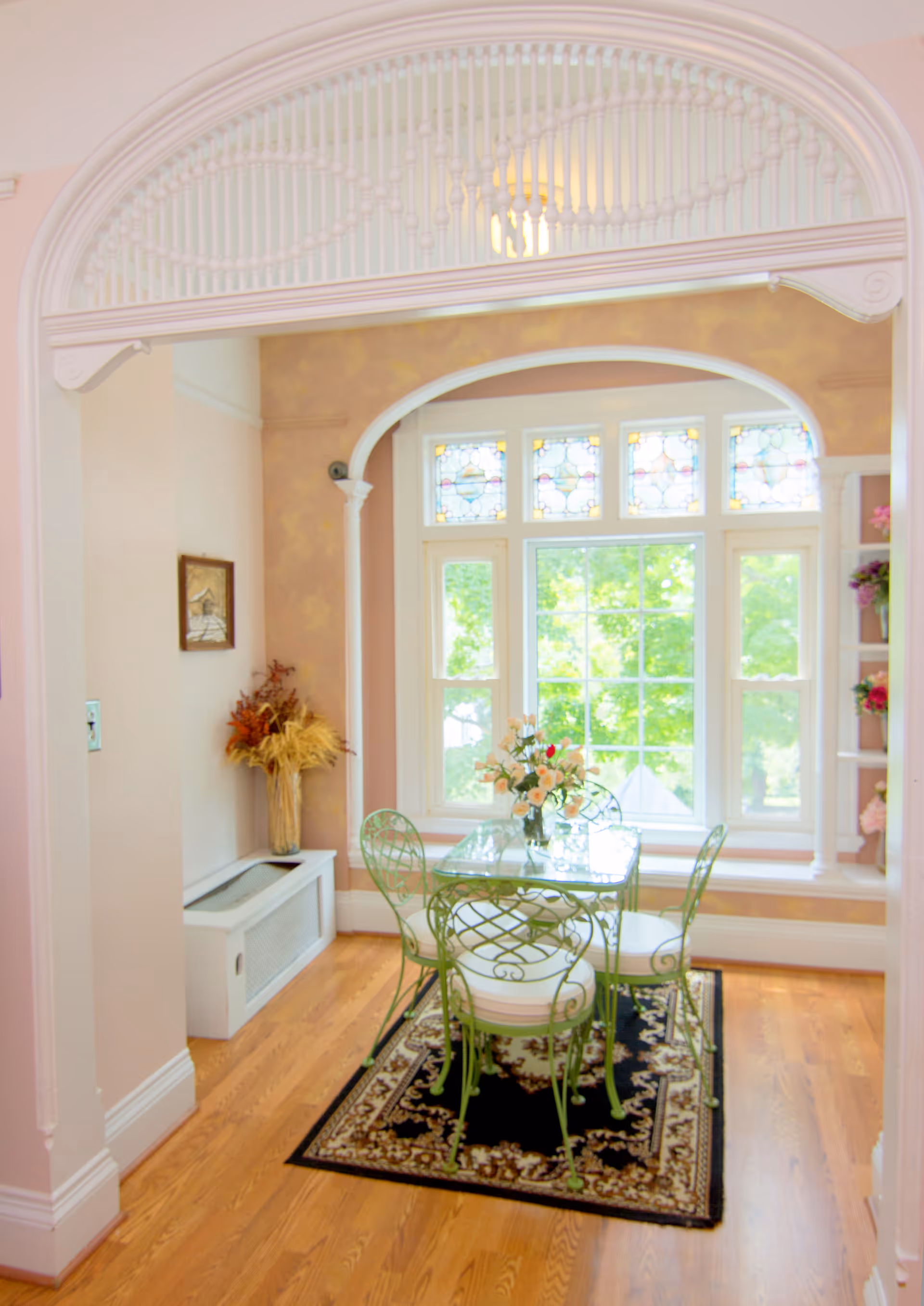 A bright dining area with a glass-top table and four green metal chairs with white cushions, placed on a decorative black and beige rug. The room features large windows with stained glass panels at the top, allowing natural light to fill the space. There is a floral arrangement on the table and dried flowers in a vase on a white radiator cover against the wall. The walls are painted in soft pastel colors with an arched doorway and decorative trim.