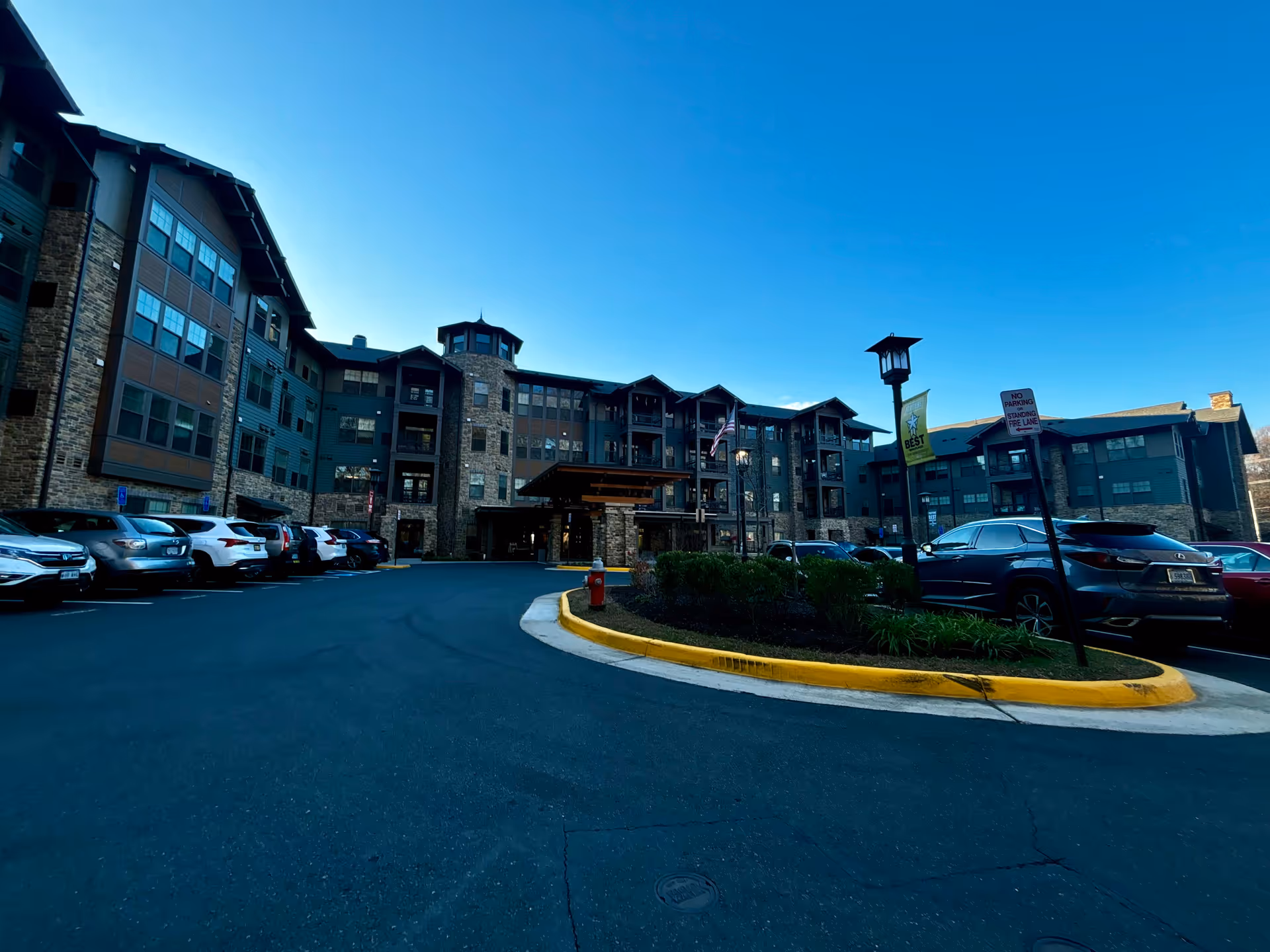 Front exterior of a multi-story senior living building with a covered entrance, parked cars, and a curved driveway under a clear blue sky.