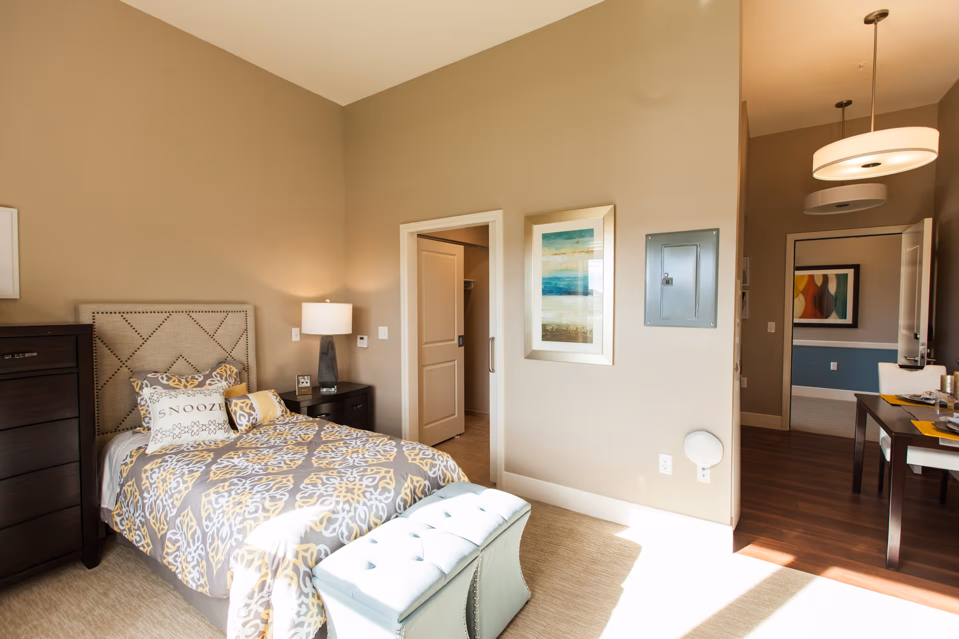 A bright bedroom with a single bed featuring a patterned gray and yellow comforter and pillows, including one with the word 'SNOOZE'. Next to the bed is a dark wooden nightstand with a lamp and an alarm clock. A matching dark wooden dresser is on the left. There is a light blue tufted bench at the foot of the bed. The walls are beige with a framed abstract painting. An open door leads to another room with a desk and chairs, and a hallway with another piece of artwork is visible in the background.