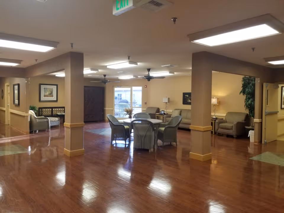 A spacious common area in a nursing and rehabilitation facility with polished wooden floors, beige walls, and ceiling lights. The room features several seating arrangements including a round table with chairs in the center, sofas, armchairs, side tables with lamps, and framed artwork on the walls. There are ceiling fans and an exit sign visible on the ceiling.