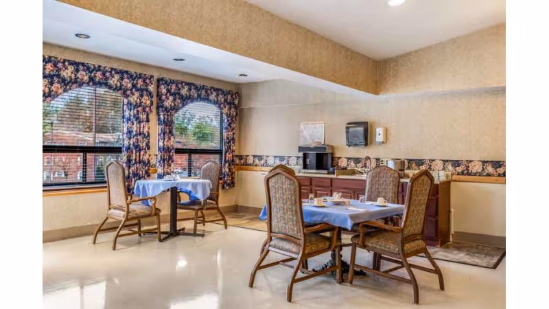A small dining area in a senior living facility with two tables covered with blue tablecloths, each surrounded by four wooden chairs with cushioned seats. The room has large windows with floral curtains, beige walls with floral wallpaper border, and a counter with a microwave, paper towel dispenser, and other items.