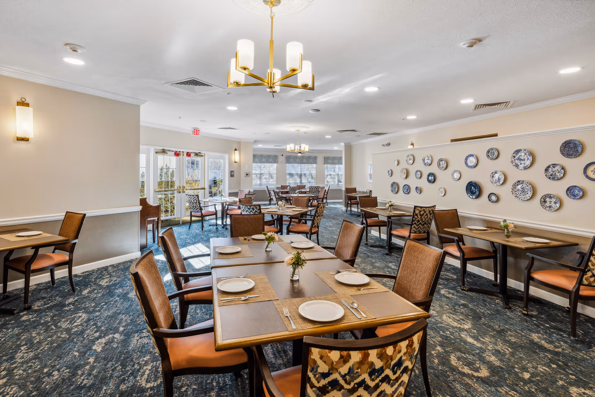 A bright and spacious dining room with multiple wooden tables set with plates, utensils, and small flower vases. The room features patterned carpet flooring, cushioned chairs with wooden frames, and decorative plates mounted on one wall. Large windows and glass doors allow natural light to fill the space, complemented by ceiling lights and wall sconces.