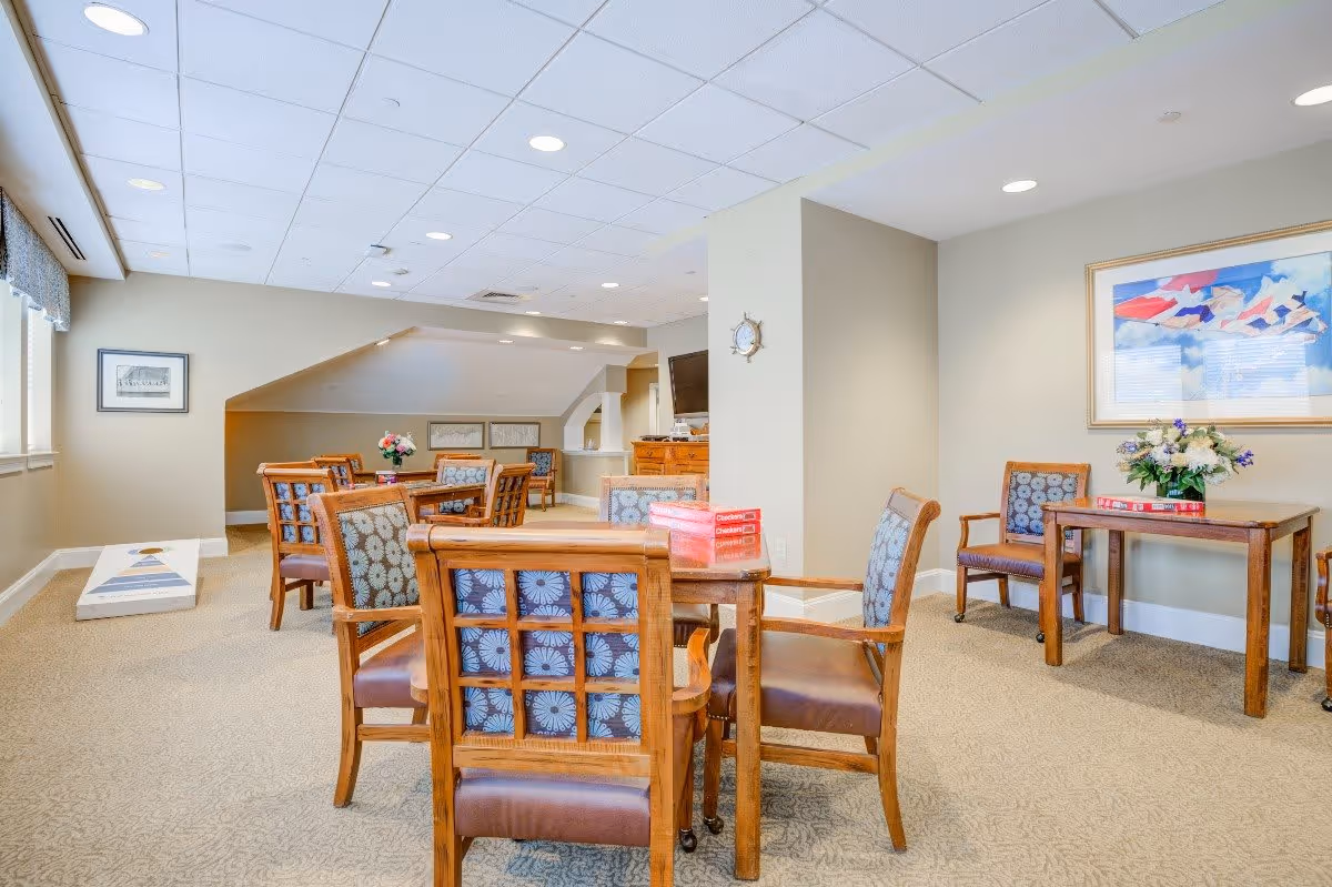 A well-lit common area with multiple wooden tables and chairs featuring patterned cushions. The room has beige walls and carpeted floors. There are framed pictures on the walls, a flower arrangement on one table, and a cornhole game set up near the window. The ceiling has recessed lighting.