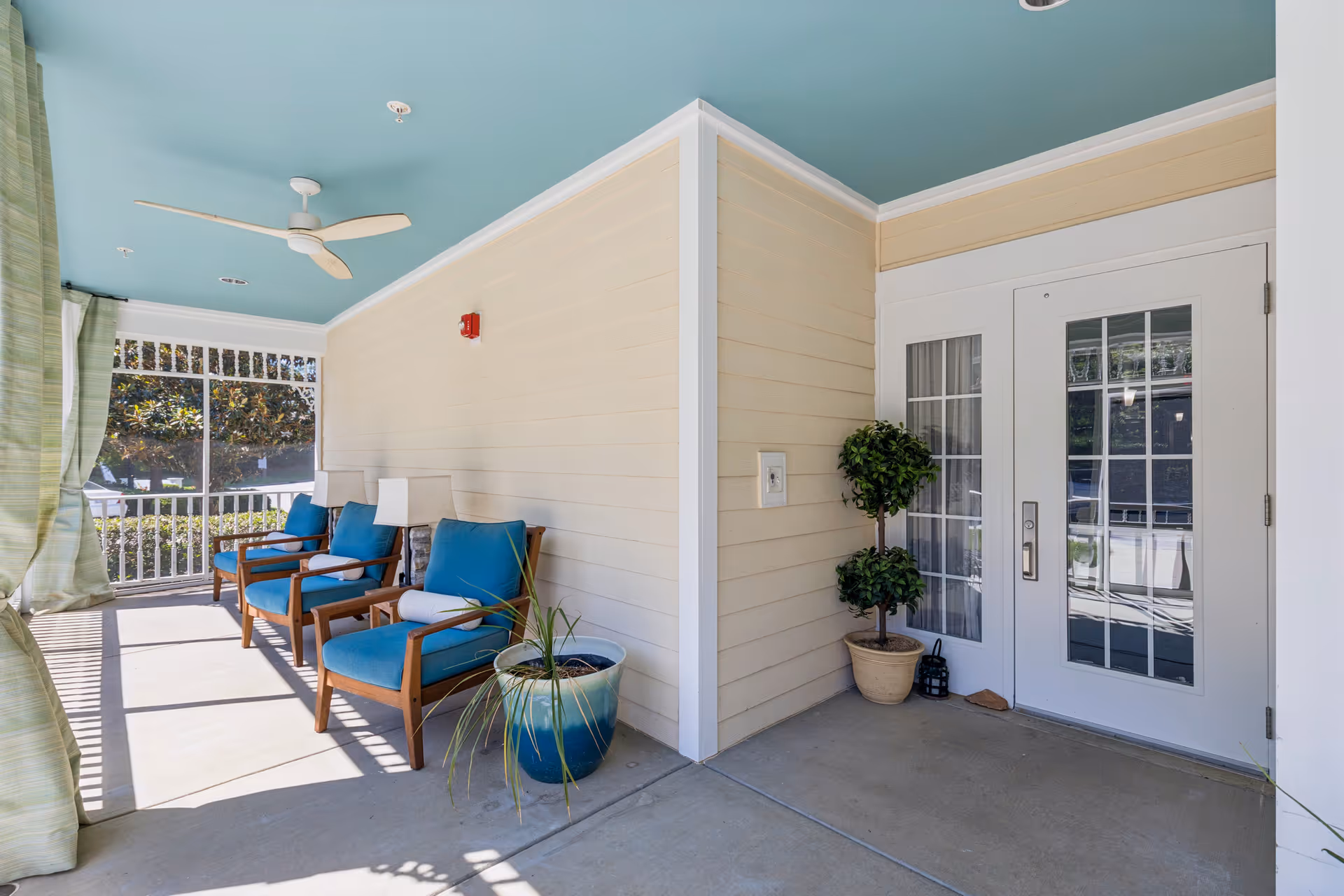 A covered outdoor porch area with three wooden chairs featuring blue cushions and white bolster pillows. There are two potted plants, one with a tall green plant in a blue and white pot and another with a small tree in a beige pot. The porch has a light blue ceiling with a white ceiling fan, cream-colored walls, and a white door with glass panels leading inside. Green curtains hang on the left side, and sunlight casts shadows on the floor.