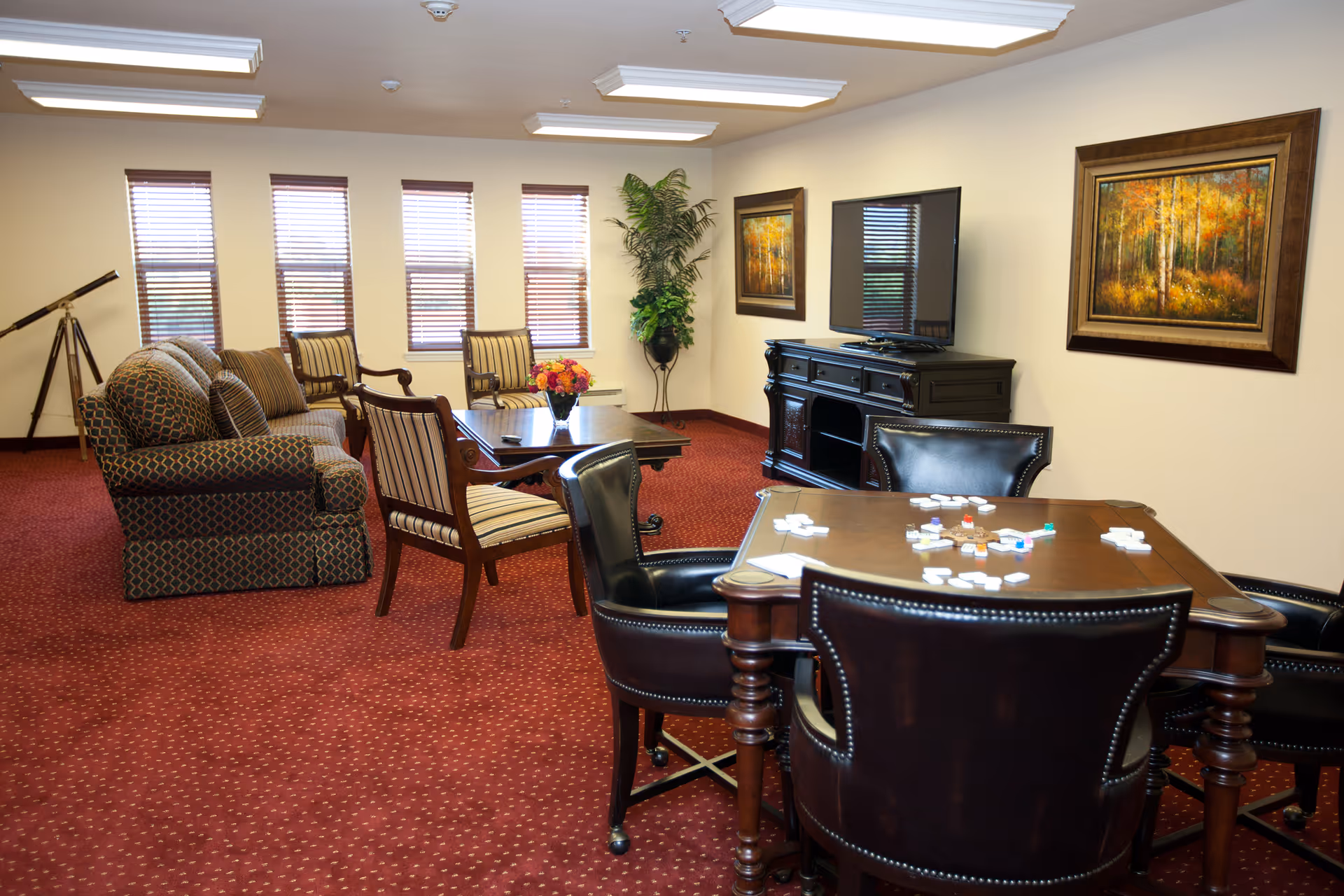 A cozy senior living common room with a red carpet, a patterned sofa, striped armchairs, a wooden coffee table with a flower vase, a TV on a dark wooden stand, and a card table with chairs and game pieces. The room has several windows with blinds, two framed paintings on the walls, and a telescope in the corner.