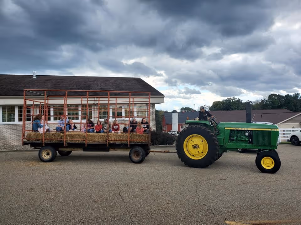 A green tractor pulls a hayride wagon full of people sitting on hay bales in front of a building under cloudy skies.