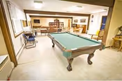 A spacious recreational room featuring a pool table in the center, surrounded by chairs and tables. The room has large windows allowing natural light, a wooden cabinet against the far wall, and a television mounted on the wall. The flooring is light-colored, and the overall ambiance is warm and inviting.