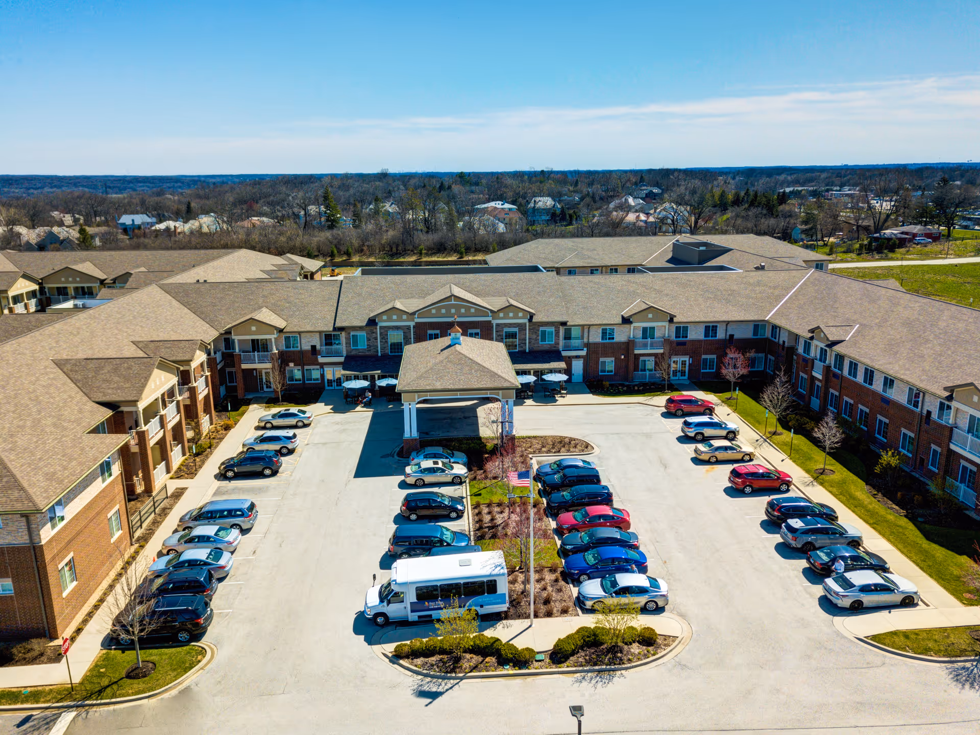 Aerial view of a large senior living facility showing the building front with a covered drop-off, surrounding parking lot filled with cars and a shuttle bus.