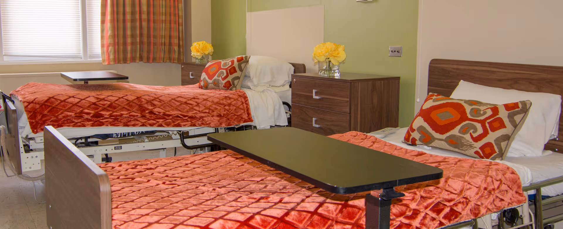 A room with two hospital-style beds, each covered with a textured orange blanket and a decorative pillow. There are two wooden nightstands between the beds, each with a vase of yellow flowers. A window with blinds and plaid curtains is visible in the background.