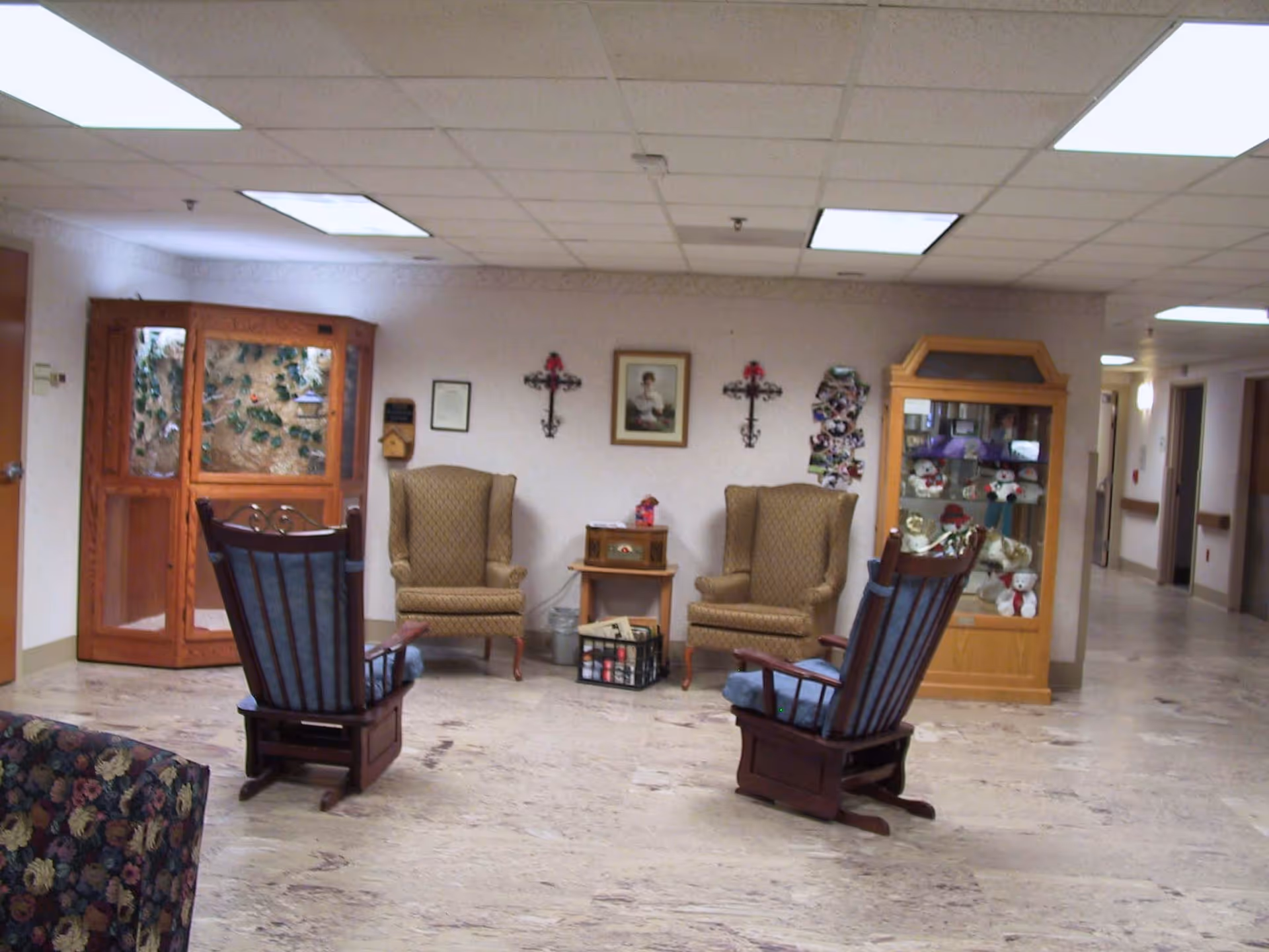 A nursing home common area with upholstered armchairs and wooden rocking chairs arranged around a small table and a display cabinet.