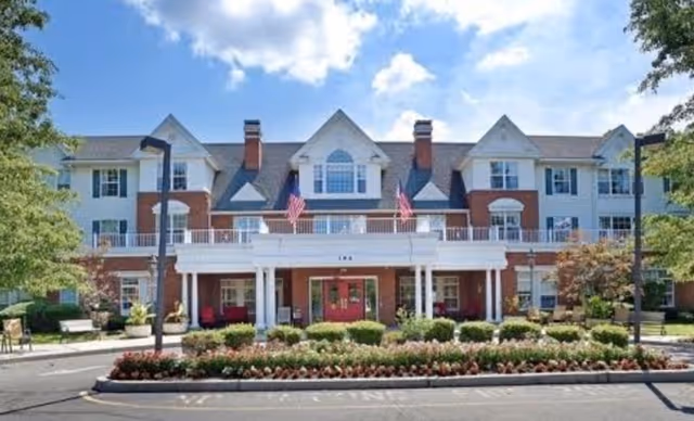 Front exterior of The New South Shore Manor, a three-story brick and siding senior living building with a covered entrance, American flags, and landscaped circular drive.