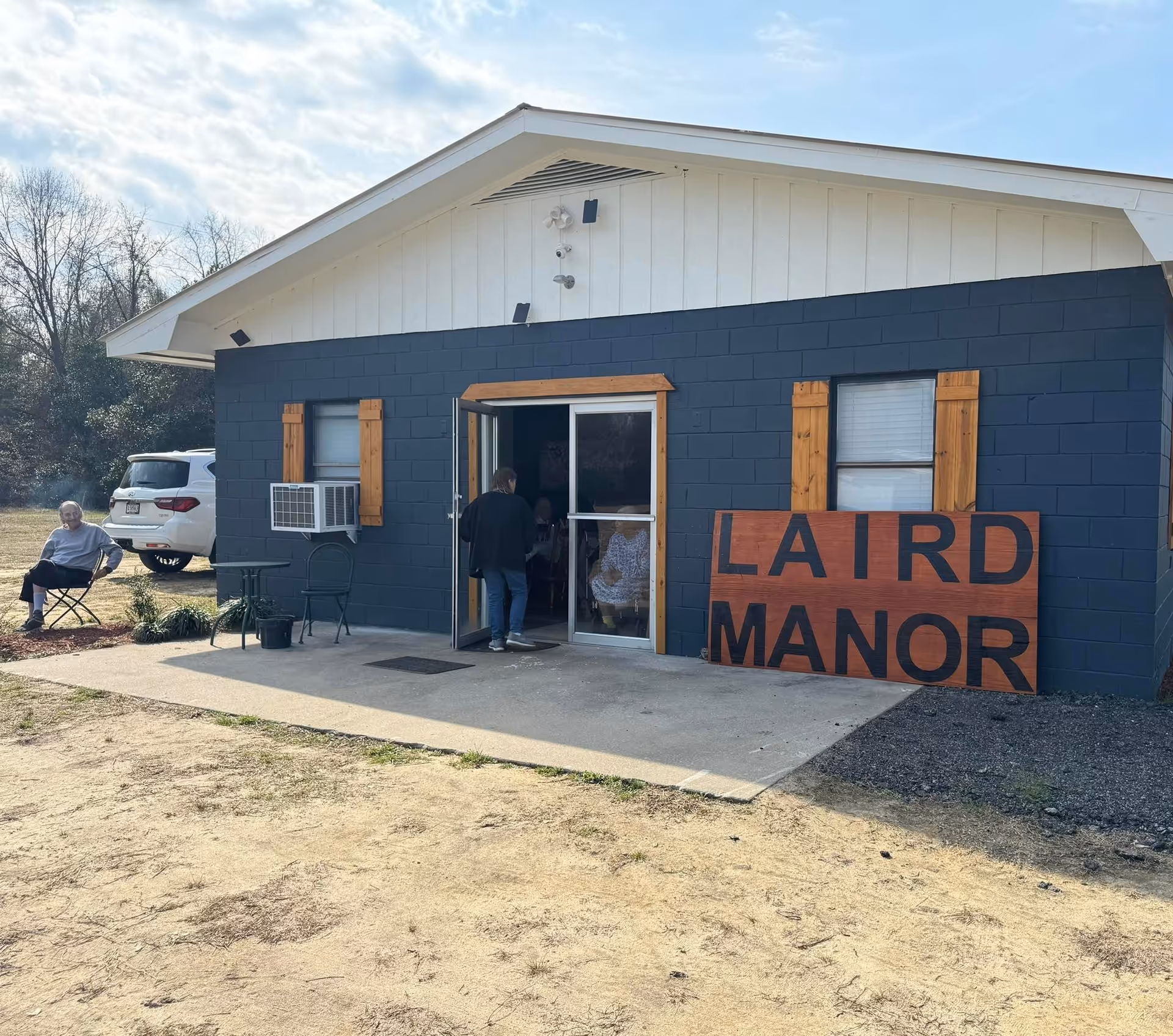 Exterior view of a small building with dark blue walls and white roof trim. There are two windows with wooden shutters and an air conditioning unit on one window. A large wooden sign leaning against the building reads 'LAIRD MANOR'. A person is entering through a glass door, and another person is sitting outside on a folding chair near a white vehicle parked on the grass. Trees and a partly cloudy sky are visible in the background.