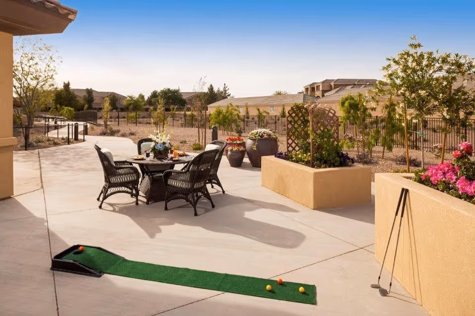 Outdoor patio area with a small round table and four wicker chairs. The table is set with flowers and drinks. There is a putting green mat with golf balls and clubs nearby. The area is surrounded by planters with flowers and small trees, and a fence encloses the space with buildings visible in the background under a clear sky.
