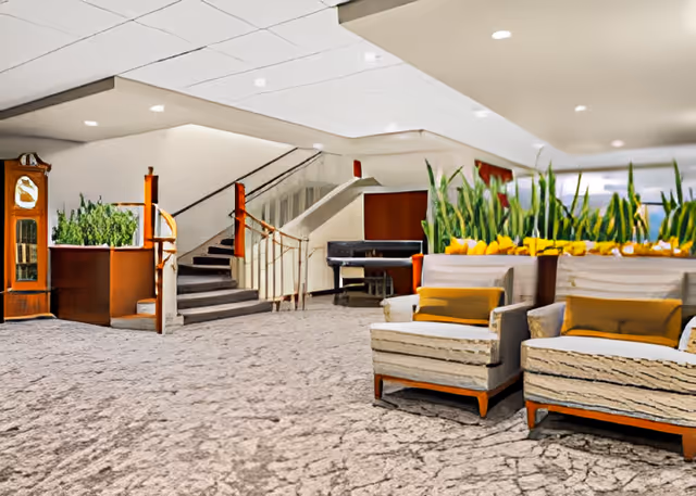 Lobby-style seating area with upholstered chairs, large planters, a staircase, a piano, and a grandfather clock.