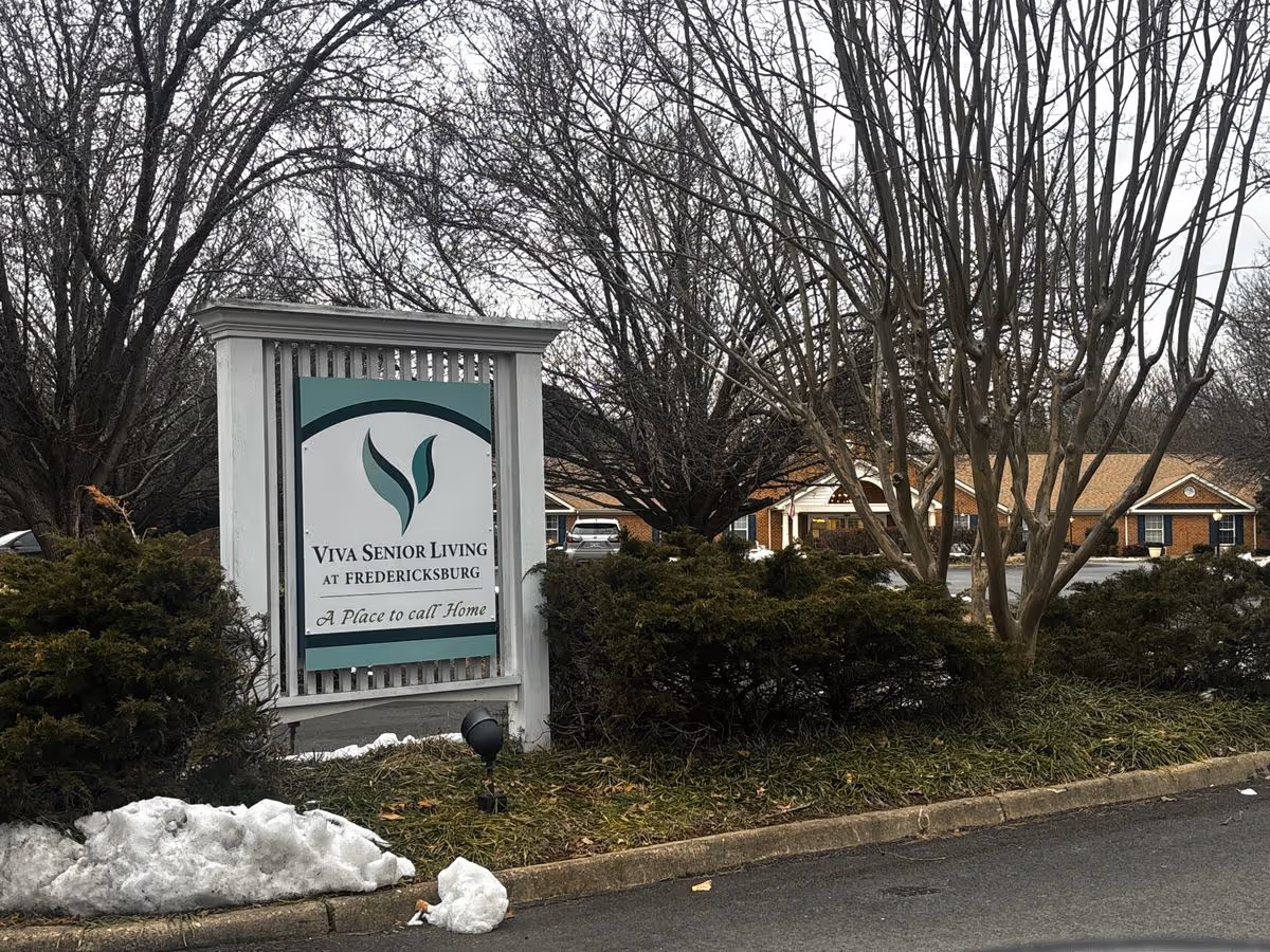 Entrance sign reading 'Viva Senior Living at Fredericksburg' set among shrubs and trees with low brick buildings in the background.