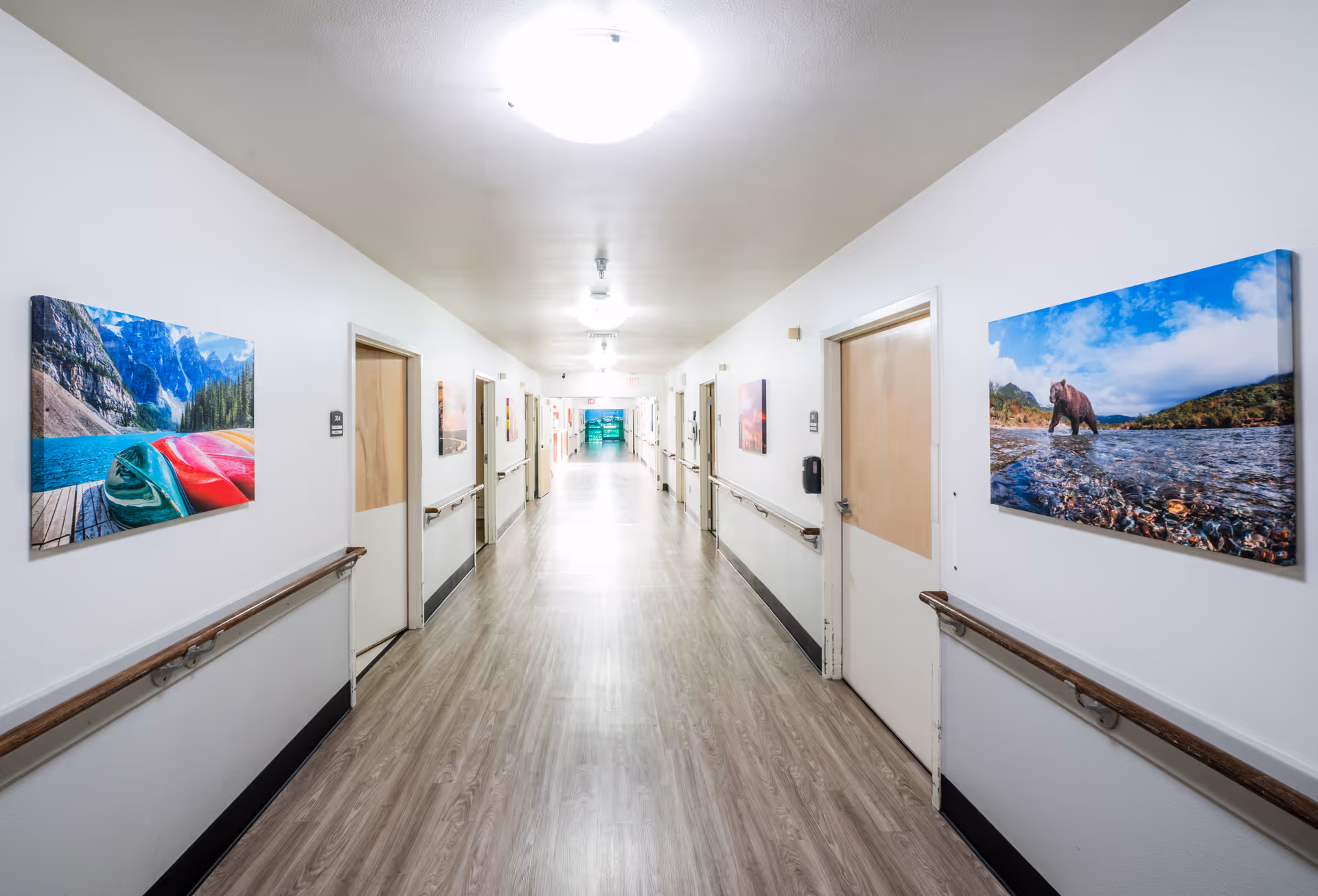A long, well-lit hallway in a senior living facility with light-colored walls and wood-patterned flooring. Several closed doors line both sides of the corridor, each with handrails beneath. Colorful landscape photographs are hung on the walls, including images of a lake with canoes and a bear in a river. The hallway extends into the distance with bright lighting overhead.