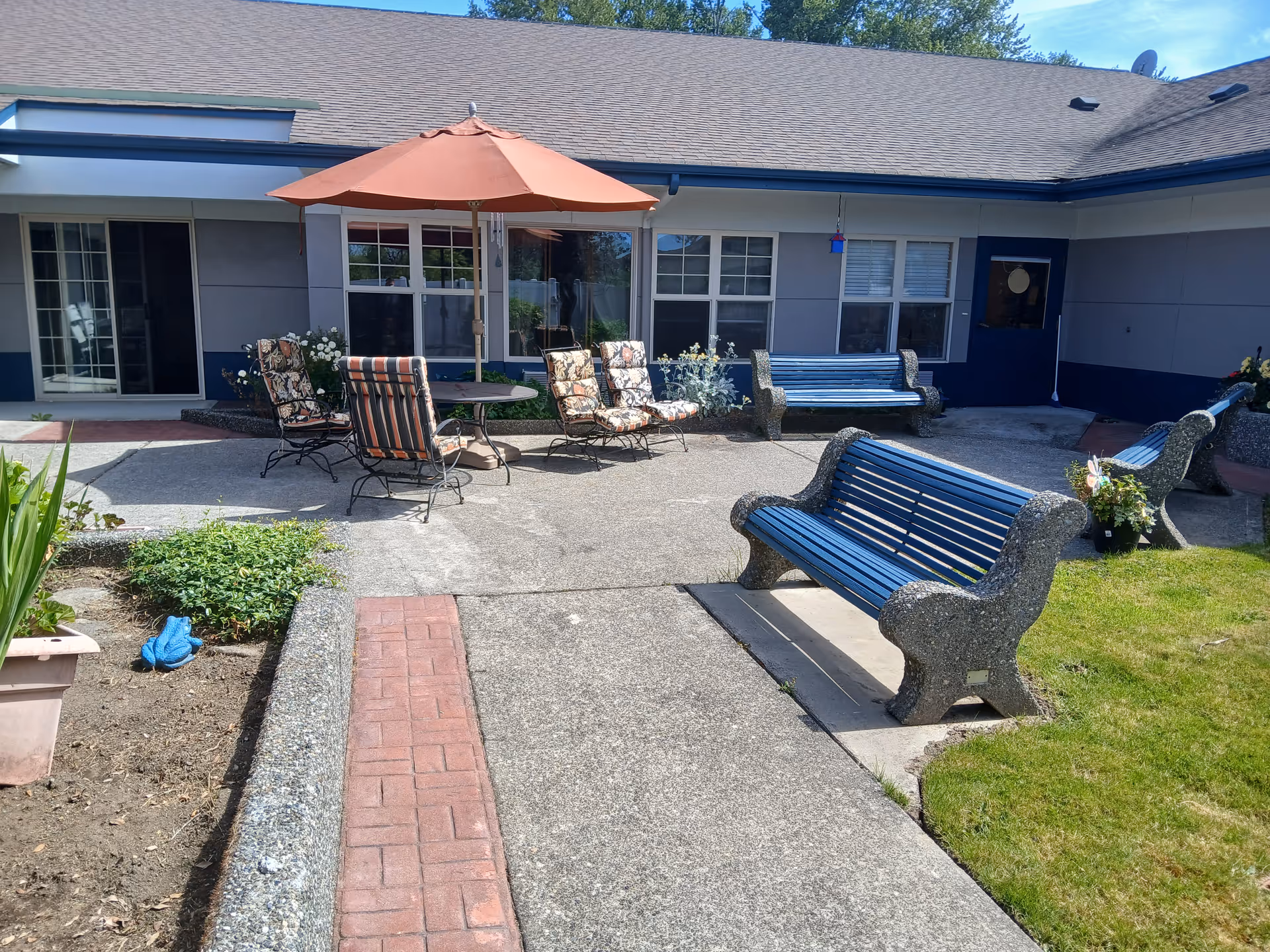 Sunny courtyard with blue benches, a patio table and chairs under an orange umbrella in front of a single-story building.