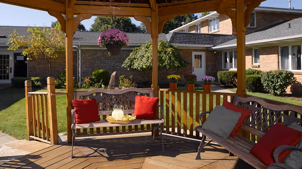 A wooden gazebo with benches featuring red and gray cushions, a pitcher of lemonade and glasses on a bench, potted flowers on the railing, and a hanging flower pot. The gazebo is situated in a grassy courtyard surrounded by a brick building with windows and doors.
