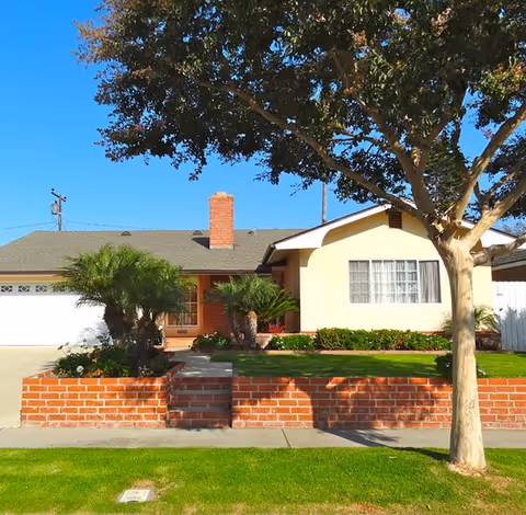 Front exterior of a single-story house with a brick planter wall, driveway and a tree in the front yard.