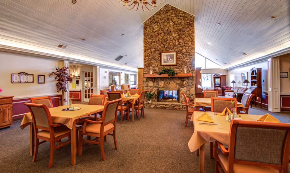 A cozy dining room with several tables covered in beige tablecloths and set with napkins and utensils. The room features a large stone fireplace in the center with a framed picture above it. There are wooden chairs with cushioned seats around the tables, and the ceiling is white with recessed lighting. The room has a warm and inviting atmosphere with additional seating and decor visible in the background.