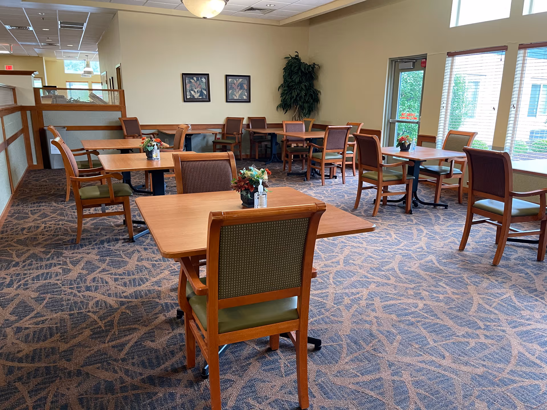 Bright communal dining room with wooden tables and chairs arranged on patterned carpet near large windows.