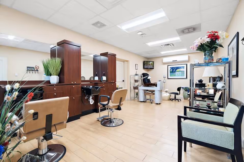 Interior view of a well-lit salon area in a senior living facility with wooden flooring, beige walls, and ceiling lights. The room features salon chairs, hair washing stations, a manicure table, and shelves with decorative items and flowers. There are framed pictures on the walls and comfortable seating for waiting.