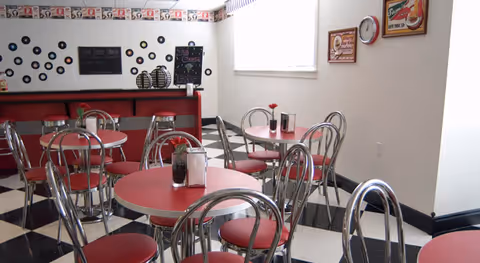 A retro-style dining area with round red tables and chrome chairs with red cushions. The floor has a black and white checkered pattern. The walls are decorated with vinyl records and vintage-style signs. There is a counter with stools along one wall and a window letting in natural light.