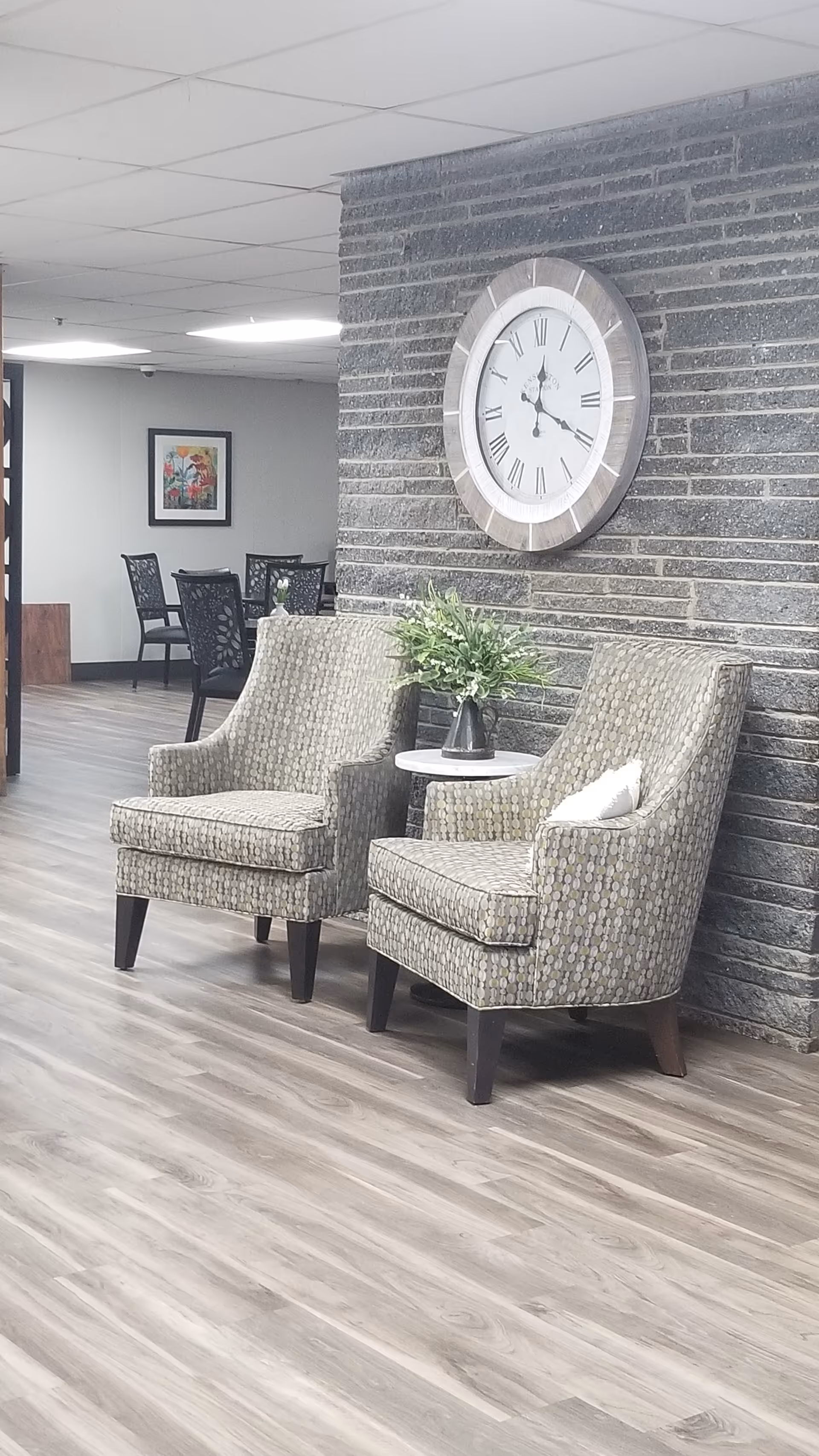 A lounge area with two patterned armchairs flanking a small table with a plant, set against a stone accent wall with a large round clock.