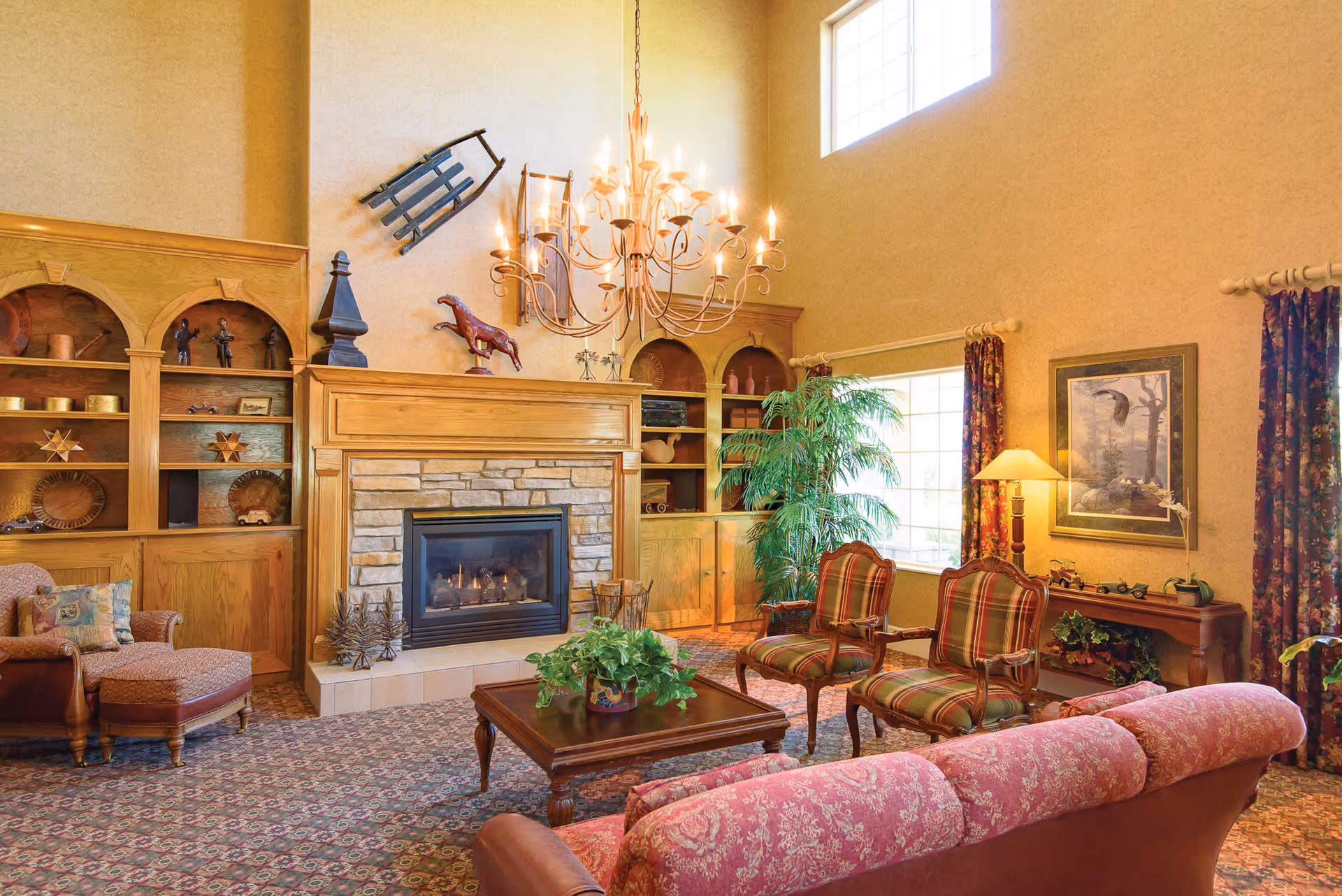A cozy living room with a stone fireplace surrounded by wooden shelves filled with decorative items. The room features a large chandelier, a tall window letting in natural light, a patterned carpet, a pink floral sofa, two plaid armchairs, a wooden coffee table with a potted plant, and a side table with a lamp and framed artwork on the wall.