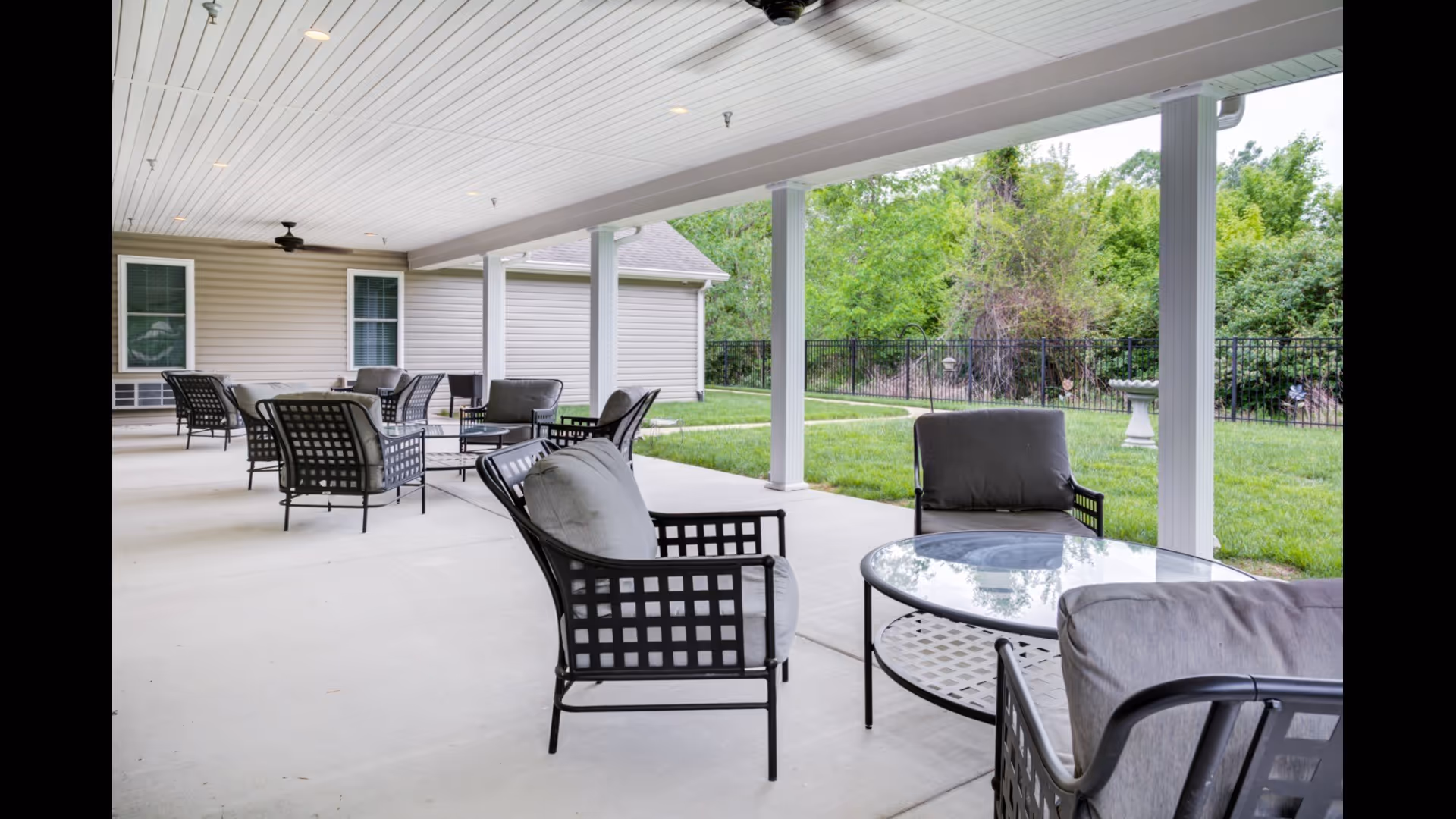 Covered outdoor patio with cushioned metal chairs and glass-top tables overlooking a grassy yard and fence.