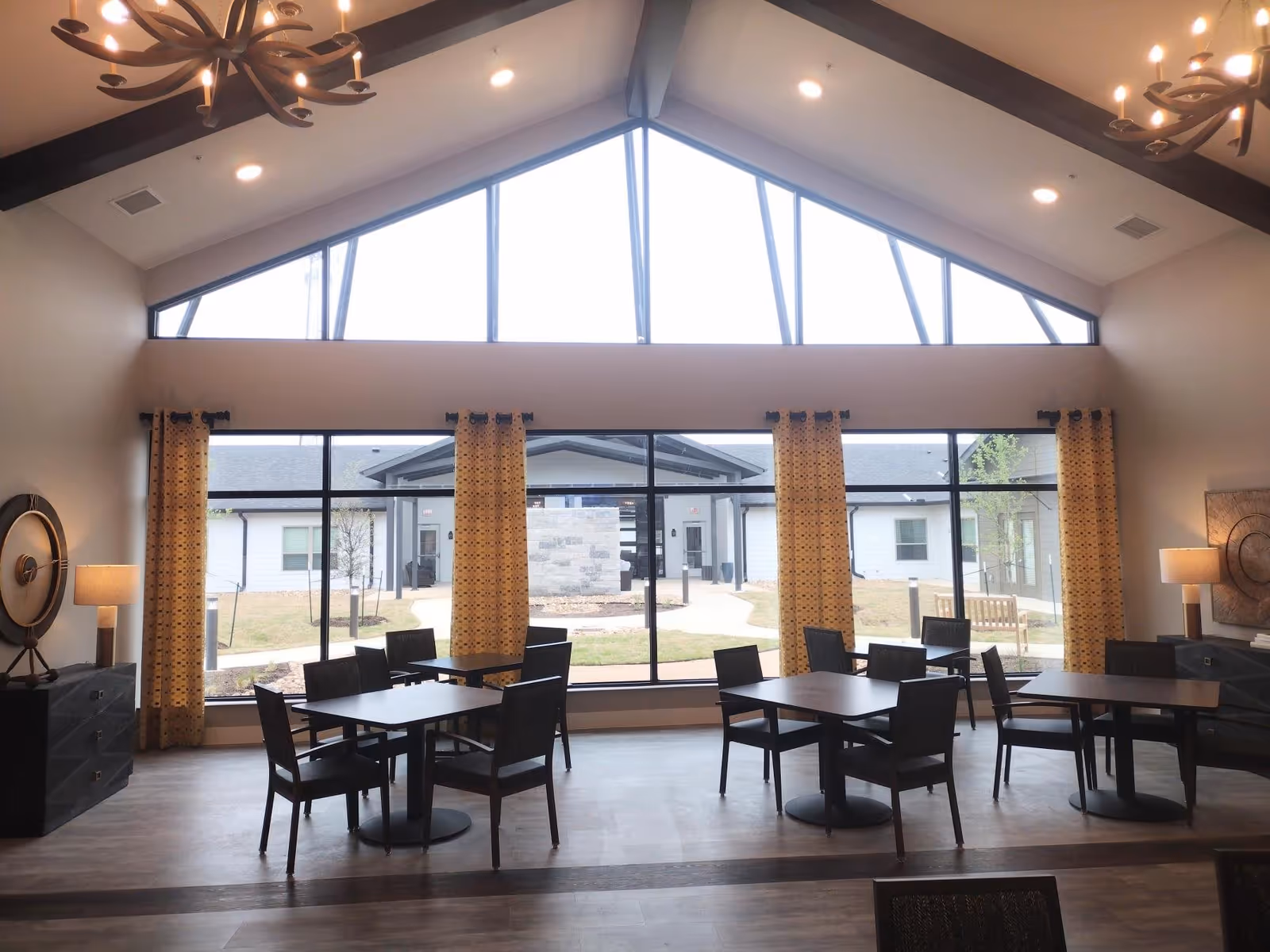 Interior view of a dining area with several dark wooden tables and chairs arranged on a wood floor. Large windows with yellow patterned curtains allow natural light to enter, showing an outdoor courtyard with a bench and a stone structure. The ceiling is vaulted with exposed beams and modern chandeliers.