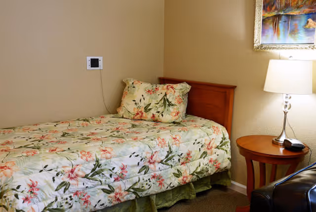 A single bed with floral bedding next to a wooden nightstand topped with a lamp and phone in a small bedroom.