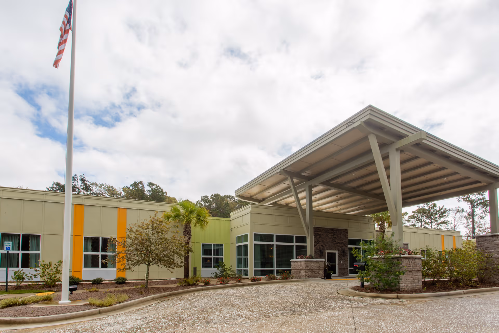 Exterior view of Ashley Gardens Assisted Living & Memory Care facility showing the entrance with a large covered drop-off area, windows, some landscaping including small trees and bushes, and an American flag on a flagpole.