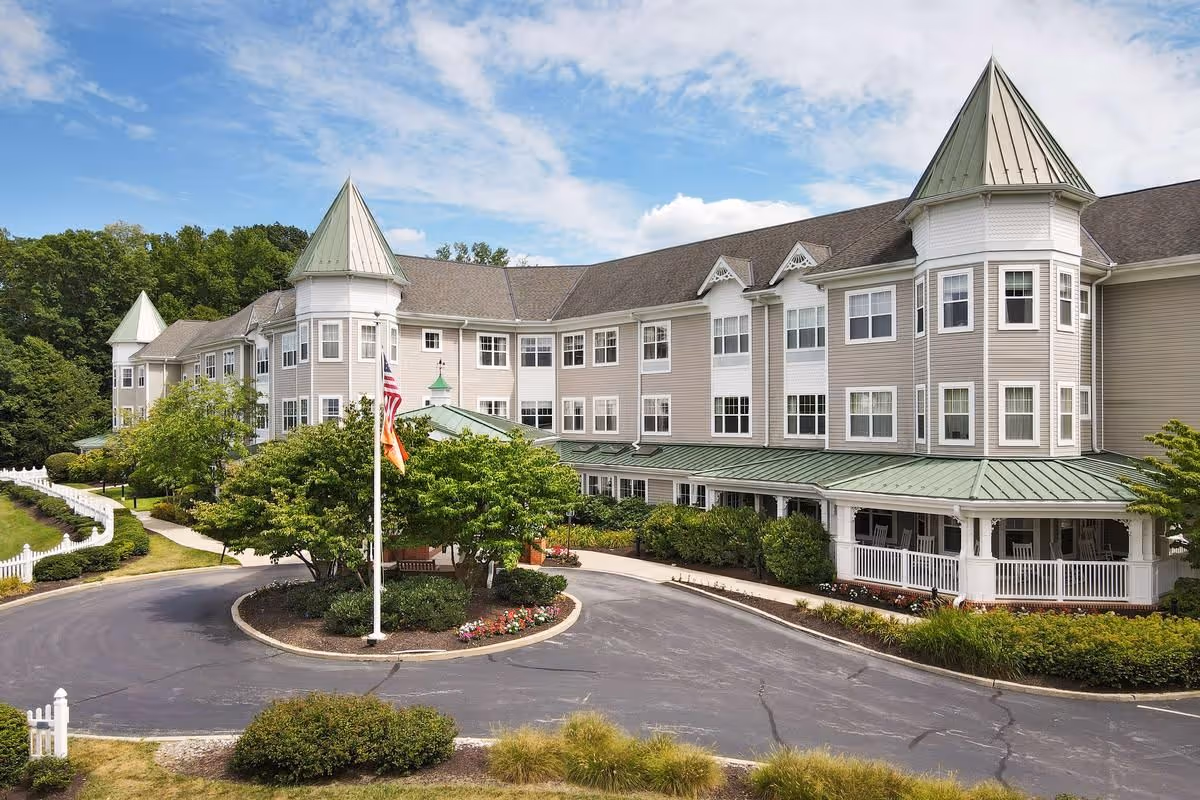 Exterior view of a large senior living facility building with beige siding, green roofs, and multiple windows. The building has two prominent turret-like structures and a circular driveway with a flagpole in the center surrounded by trees and landscaping. The sky is partly cloudy.