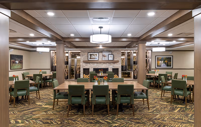 Spacious communal dining room with wooden tables, green upholstered chairs, a central fireplace, and warm overhead lighting.