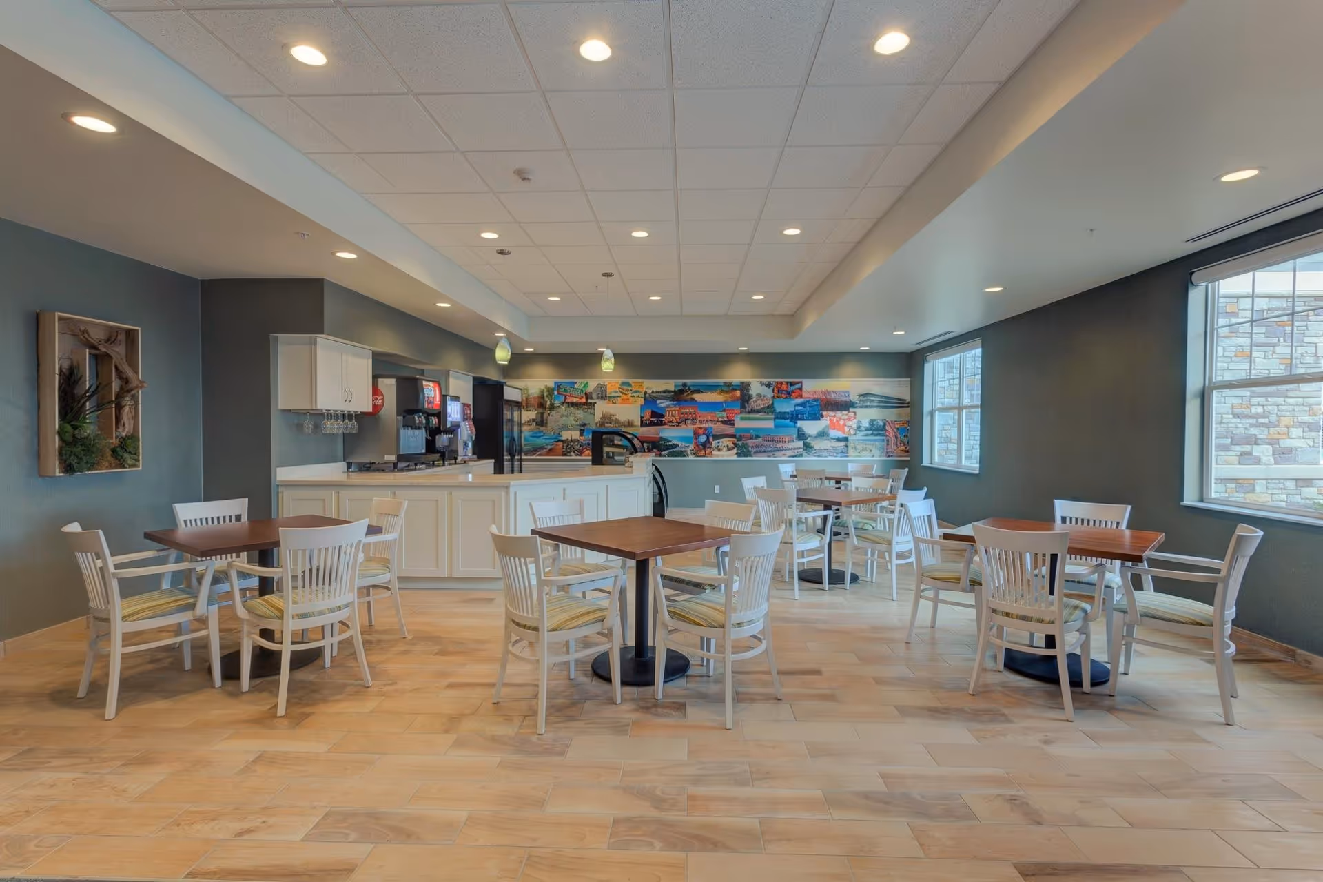 A bright dining area with several wooden tables and white chairs with striped cushions. The room features a beverage station with soda dispensers and a refrigerator along the back wall. Large windows on the right side allow natural light to enter, and a colorful mural decorates the back wall above the beverage station. The floor is light wood, and the ceiling has recessed lighting.
