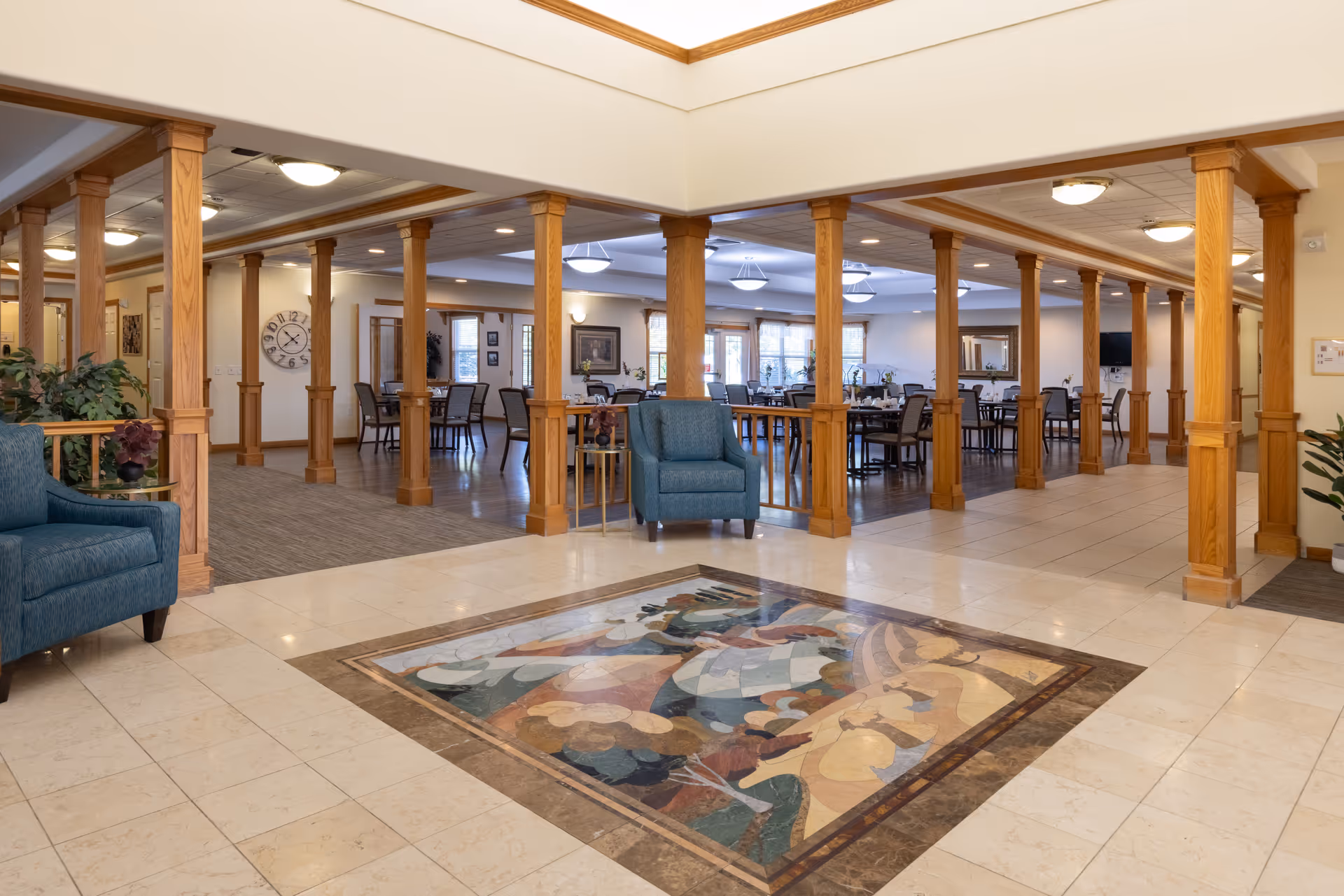Spacious interior of a senior living facility featuring a tiled floor with a colorful mosaic design in the center, wooden columns separating a seating area with blue armchairs from a dining area with multiple tables and chairs. The space is well-lit with ceiling lights and has large windows allowing natural light.